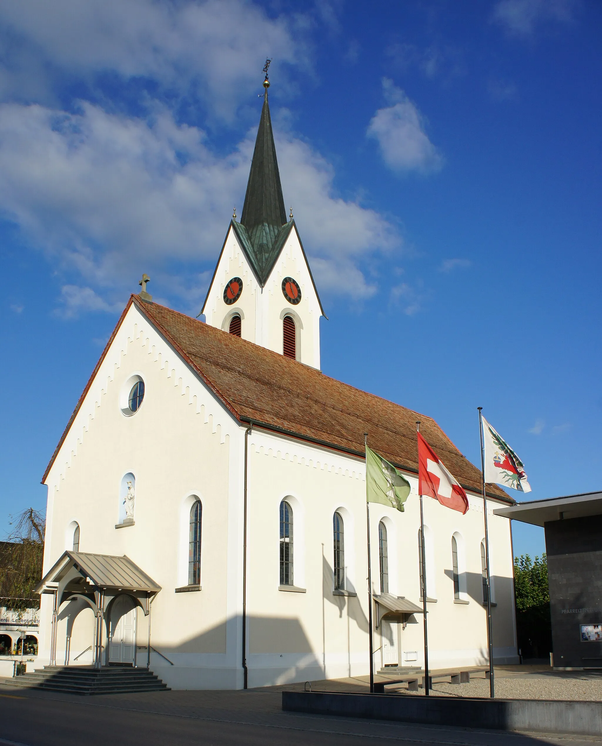 Photo showing: Die Kirche Kriessern aus südwestlicher Richtung, der mit einem Satteldach überdachte Haupteingang an der Westfassade ist vom Trottoir aus über 6 Stufen erreichbar, der mit einem kleinen Schrägdach geschützte Seiteneingang an der Südfassade ist vom Vorplatz über 2 Stufen erreichbar, der Kirchturm steht an der Nordost-Ecke, rechts im Vordergrund steht ein länglicher Brunnen parallel zur Strasse, dahinter drei Fahnenmasten (von l. nach r.; St. Galler-, Schweizer- und Kriessner-Fahne)