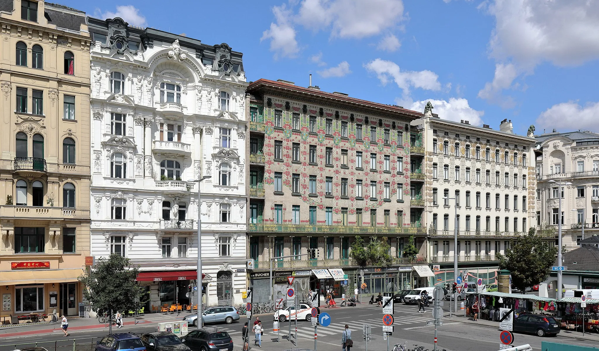 Photo showing: Die Linke Wienzeile auf der Höhe der Kettenbrücke(ngasse) im 6. Wiener Gemeindebezirk Mariahilf mit dem „Majolikahaus“ von Otto Wagner im Bildzentrum. Ganz links Haus Nr.44, Wohnhaus des Heldentenors Hermann Winkelmann (1849–1912), anschließend Haus Nr. 42, ein reich gegliedertes historistisches Wohn- und Geschäftshaus, das von 1896 bis 1897 nach Plänen von Rudolf Kmunke errichtet wurde. Im Anschluss Haus Nr. 40, auch „Majolikahaus“ bezeichnet, ein Miethaus, das von Otto Wagner 1898/1899 errichtet wurde und das Eckhaus „Köstlergasse“ mit vergoldetem secessionistischem Dekor, wiederum ein von Otto Wagner 1898/1899 errichtetes Haus und ganz rechts Haus Nr. 36, ein monumentales späthistoristisches Mietpalais, erbaut 1897 nach Plänen von Franz Ritter von Neumann. Rechts im Vordergrund die Kettenbrücke und der Naschmarkt.