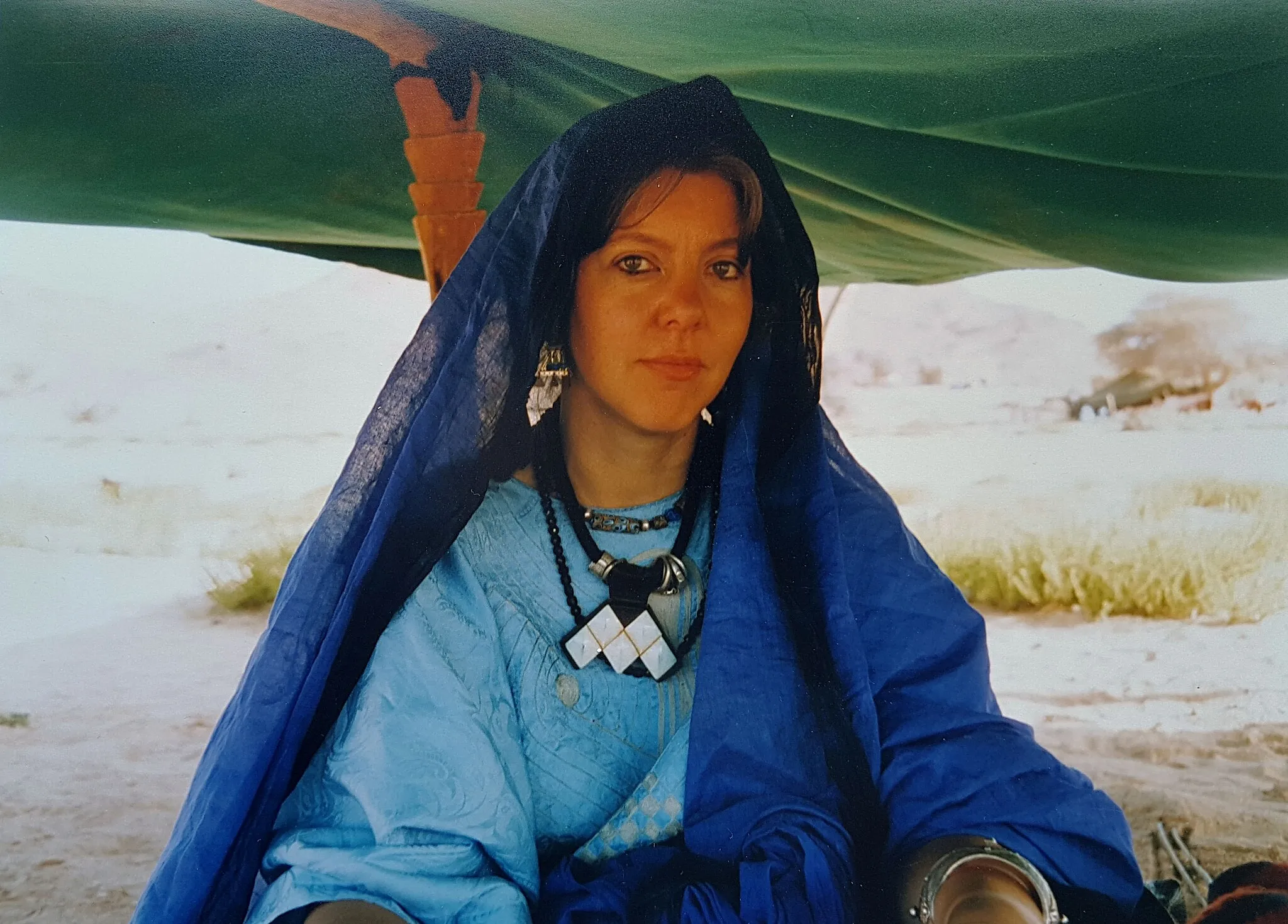 Photo showing: Dutch social antropologist and artist Angeline van Achterberg (1949-2020) is wearing Tuareg jewelry in Algeria: a silver bracelet with small knobs, three necklaces, one of which with a large geometric metal ornament (M-shaped pectoral pendant) and elaborate silver ear hangers. She is dressed in light blue attire under a darker blue transparent veil and she is sitting under a green cloth supported by a wooden beam. In the background a landscape with yellow grass and a tree.