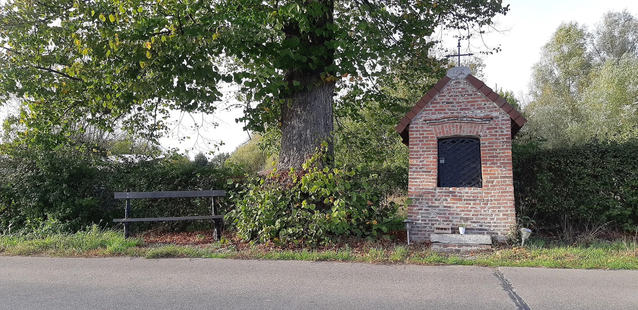 Photo showing: Pijlerkapel gewijd aan Onze-Lieve-Vrouw van Zeven Weeën, geflankeerd door een linde (Tilia spec.) als kapelboom en voorafgegaan door een stenen knielbank. Verankerde baksteenbouw onder zadeldak bedekt met tegelpannen en bekroond met een gesmeed ijzeren kruis. Licht getoogde nis onder strek en getrapte gebogen druiplijst. De nis wordt afgesloten door gesmeed ijzeren traliewerk en bevat een gepolychromeerd Onze-Lieve-Vrouwebeeld met Kind op een gemetste sokkel. (naar: https://inventaris.onroerenderfgoed.be/erfgoedobjecten/308335)