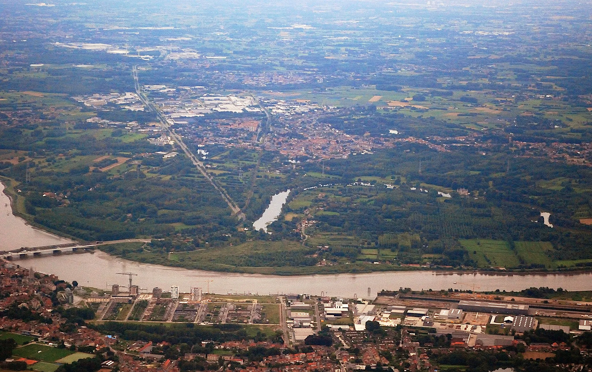 Photo showing: Aerial photograph of the Flemish town of Bornem viewed from the northeast. Near the bottom is the town of Temse on the left bank of the Scheldt, with Temse bridge and the historic crane of the former Boel shipyard. In the background behind Bornem is Puurs and then, slightly to the left, Kalfort. Nikon D60 f=55mm f/5.6 at 1/640s ISO 800. Contrast and colours enhanced using Nikon NX Studio 1.3.2. Framed and sharpened using GIMP 2.10.34.