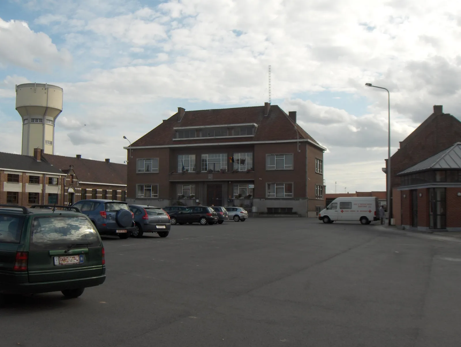 Photo showing: Basècles, Ortsteil der Gemeinde Beloeil im Hennegau (Belgien), Ortsmitte mit Asphaltparkplatz aus den 80er Jahren des 20. Jh.s, ehemaliges Rathaus im Hintergrund, heute örtliches Sozialamt.