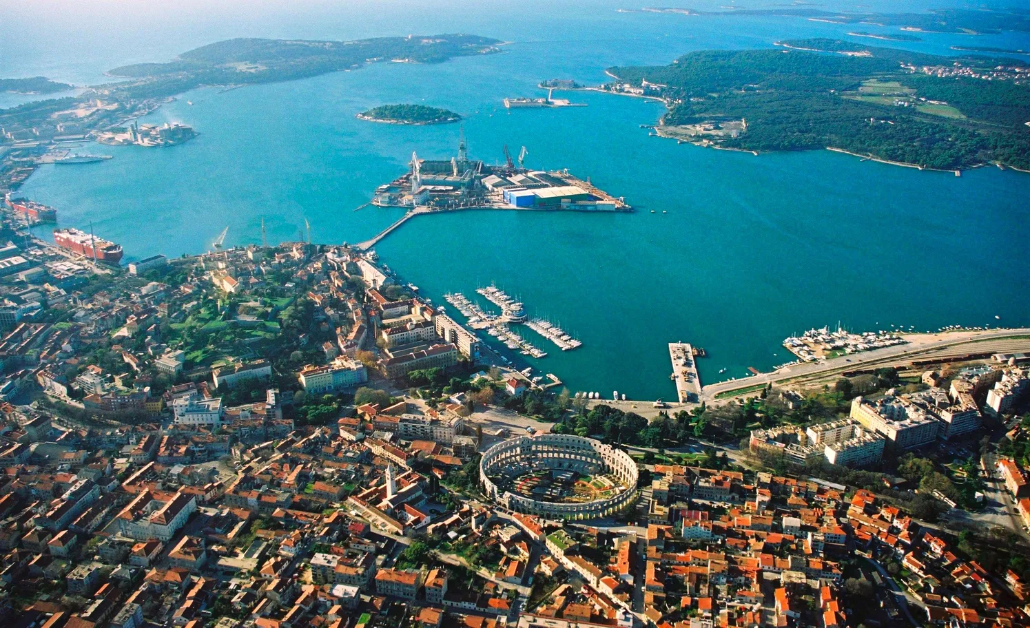 Photo showing: Aerial view of Pula, Croatia. The most notable object, on the bottom of the photo, is the Arena Amphiteathre. Left of it is the hill around which lies the Old Town. The wedge-shaped island in the middle of the bay is Uljanik, part of the shipyard. On the far top right the Brijuni islands (a National Park) can be seen.