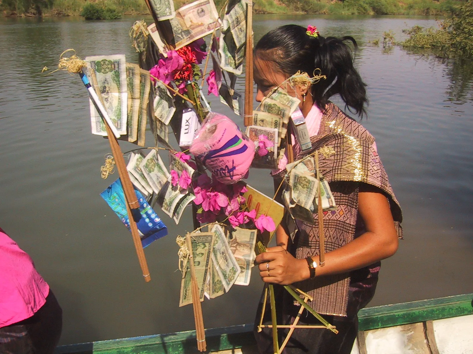 Photo showing: Beautifully dressed lady carrying a buddhist money tree in Laos. Among the symbolic leaves there are banknotes, handmade incense, toilet paper, pen, washing powder, soap, book, cigarettes, and flowers.