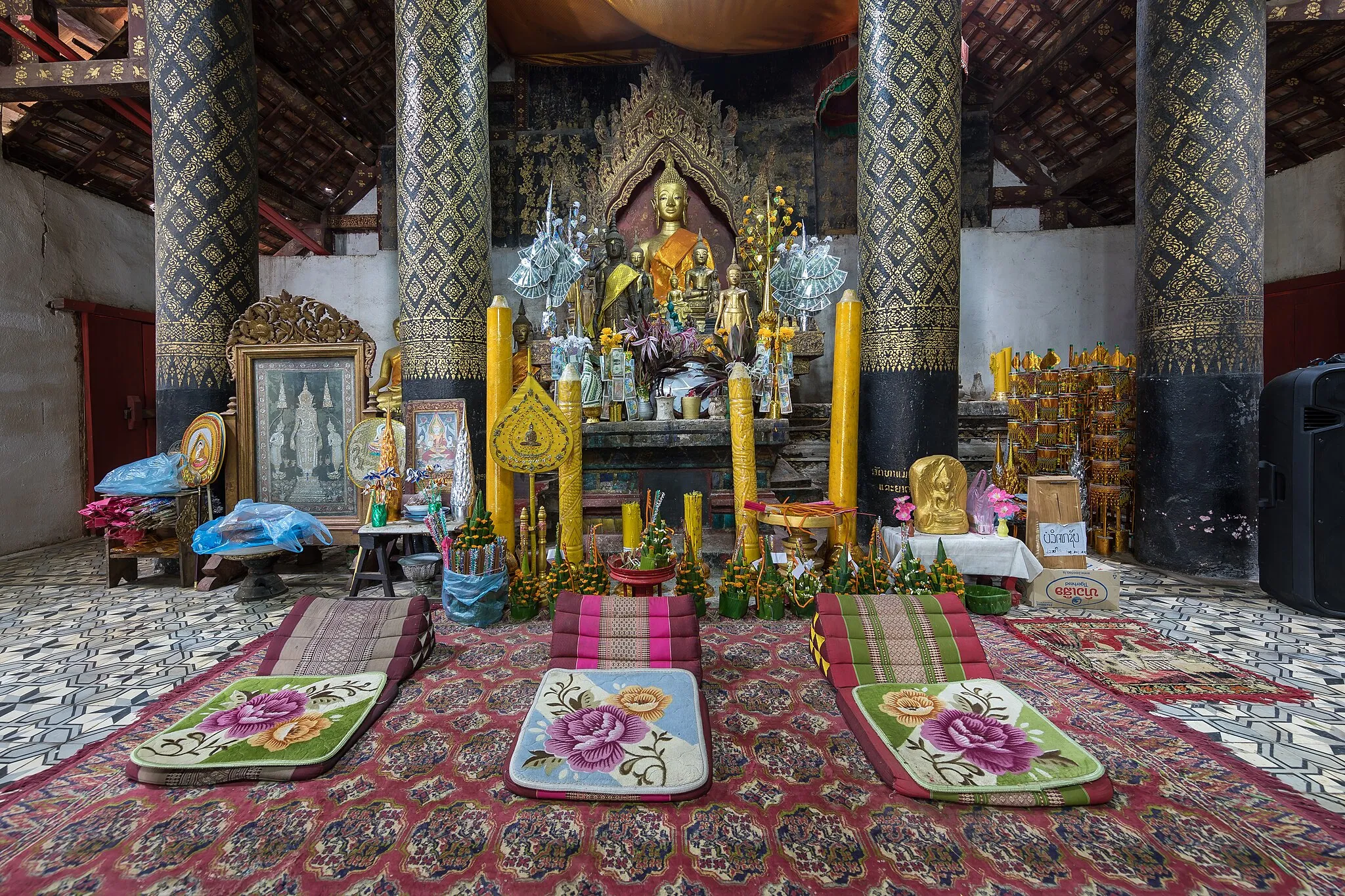 Photo showing: Praying area inside the temple Wat Jom Phet of Luang Prabang (Laos), with painted pillars, a golden statue of the Buddha, big yellow candles, and three triangle prayer cushions