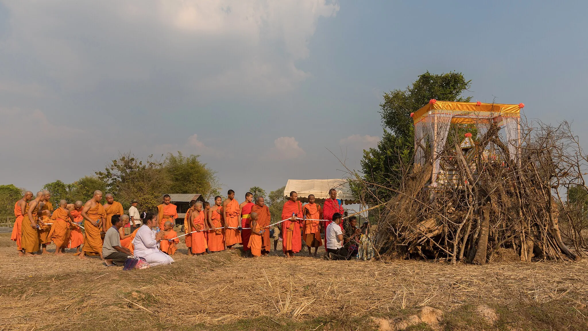 Photo showing: Ritual procession of Buddhist monks in front of a coffin over a pyre before lighting the fire, during funerals in the countryside of Don Det, Laos.
Holding a rope linked to the coffin is a ritual called chungsob (ຈູງສົບ in lao) in relation with the soul of the deceased. This rope will not be incinerated and may be kept by the monks.

Traditionally, cotton strings play an important role in ceremonies in Laos, like in the baci, and can be used in various ways at diverse occasions. In front of the coffin, there is a Buddhist money tree, religious item with authentic banknotes, an offering to the monks.