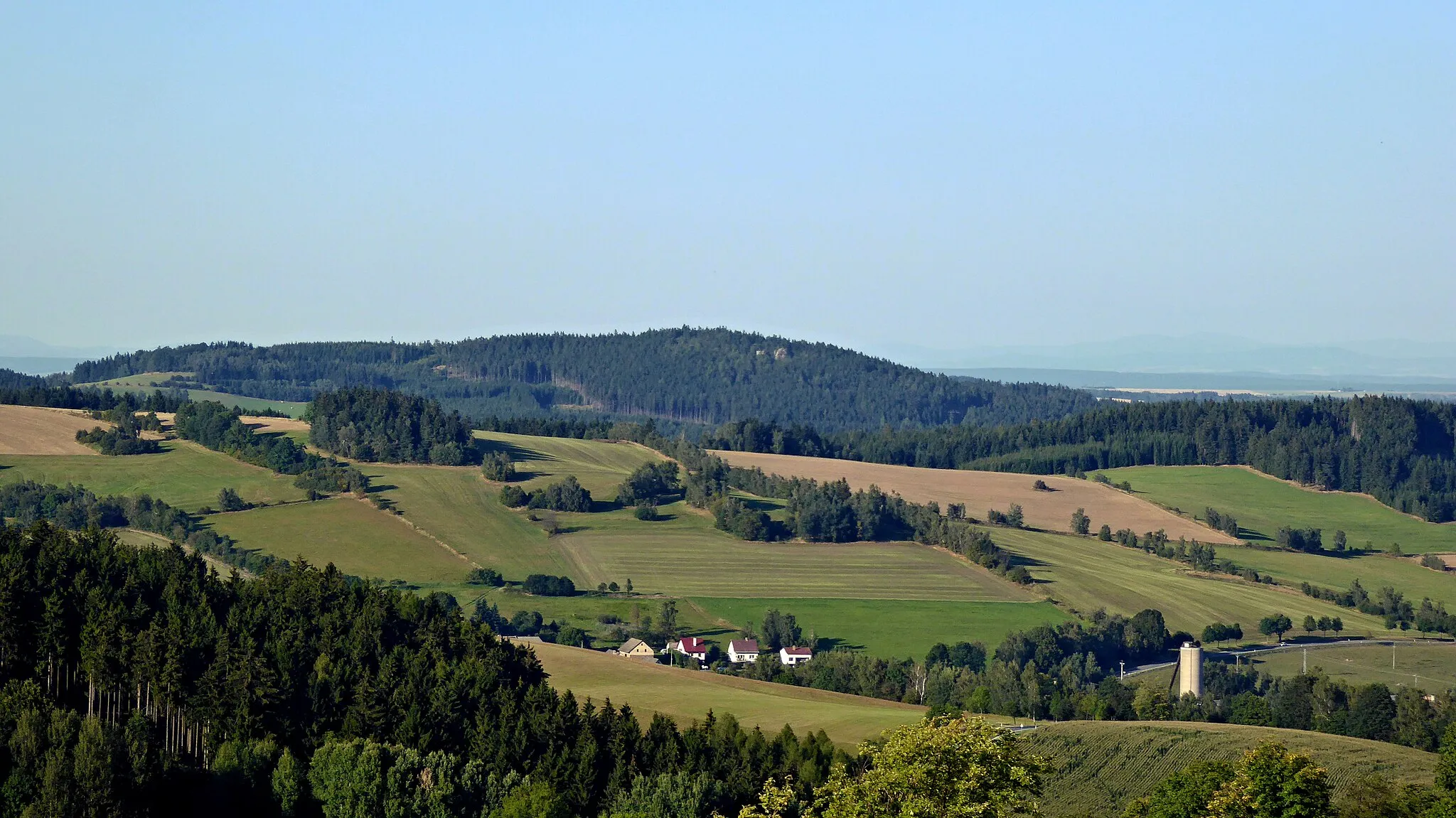Photo showing: Shape or appearance of earth surface, exactly the face of Earth, in one word georelief in of part of the "Pohledeckoskalská" Highlands. View from the slope of the "Kamenice" hill (780 m above sea level) in the northeast direction to the peak of "Prosíčka" (739.4 m above sea level), in the middle photo. Photo-location: Czechia, "Vysočina" Region, village "Roženecké Paseky" (part of municipality "Věcov"), "Pohledeckoskalská" Highlands (30°). The rocks modeling surface forms of the earth's crust, is part of the litosphere of the Earth. Georelief is very complicated, the upper surface forms soil cover (pedosphere) of different thickness. In many places, rock outputs of varying sizes and heights appear. The georelief is broken down by height in to two basic types, are it lowlands or highlands and also subtypes, for example a plains, a land knoll hills (czech landscape of small hills, hilly country), a highlands, a mountains and big mountains. In regional divide are types of georelief localized into territorial geomorphological units. In the Czech Republic there is a division: province, system, sub-system, whole (in territory complex), part of whole (of complex) and the district. The "Pohledeckoskalská" Highland is a type of rugged highland at altitudes of 395-822 metres forming a geomorphological district.