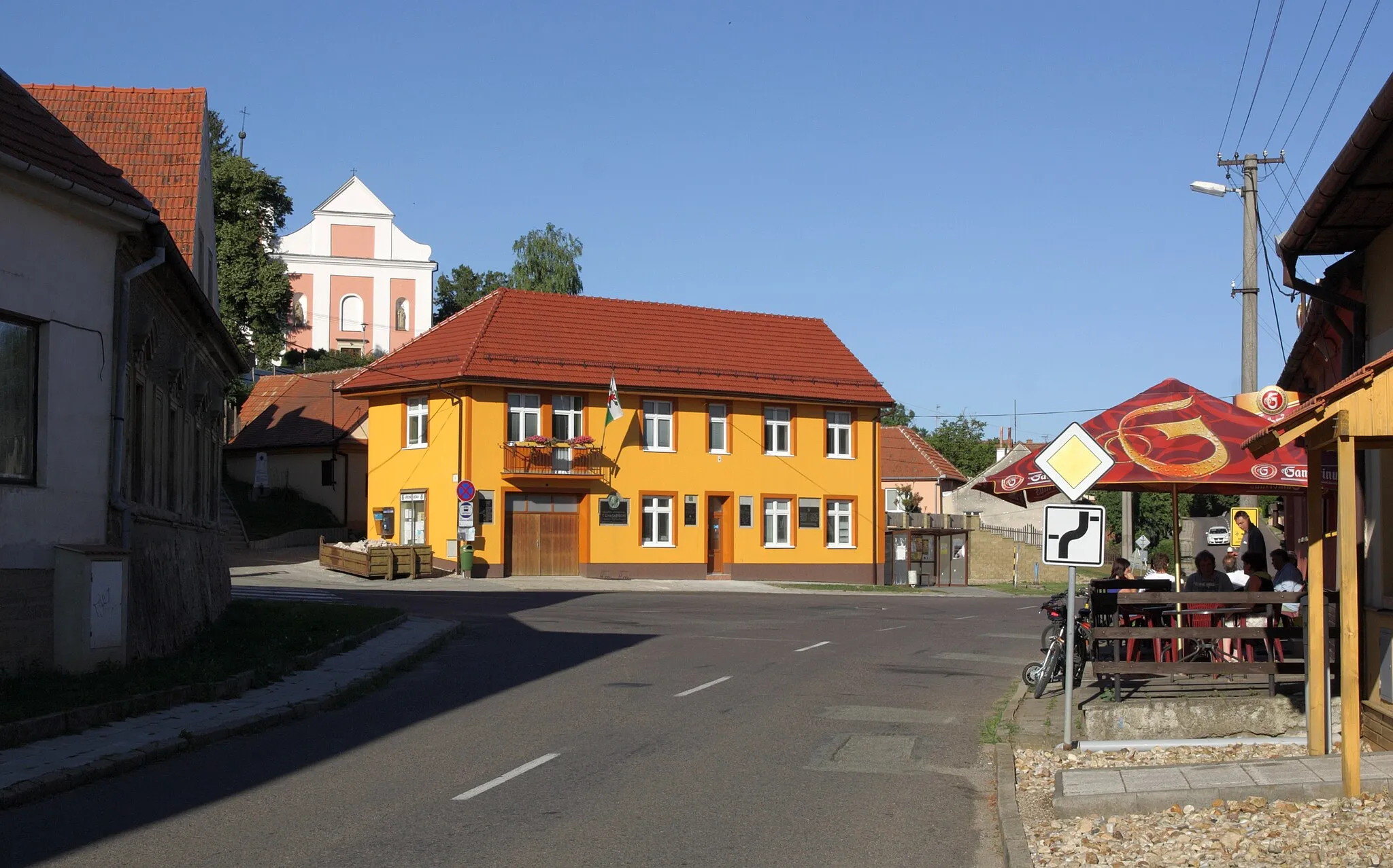 Photo showing: Velké Hostěrádky - náves s obecním úřadem, v pozadí kostel svatého Šebestiána, Rocha a Rozálie. Po levé straně silnice bývalý zájezdní hostinec.