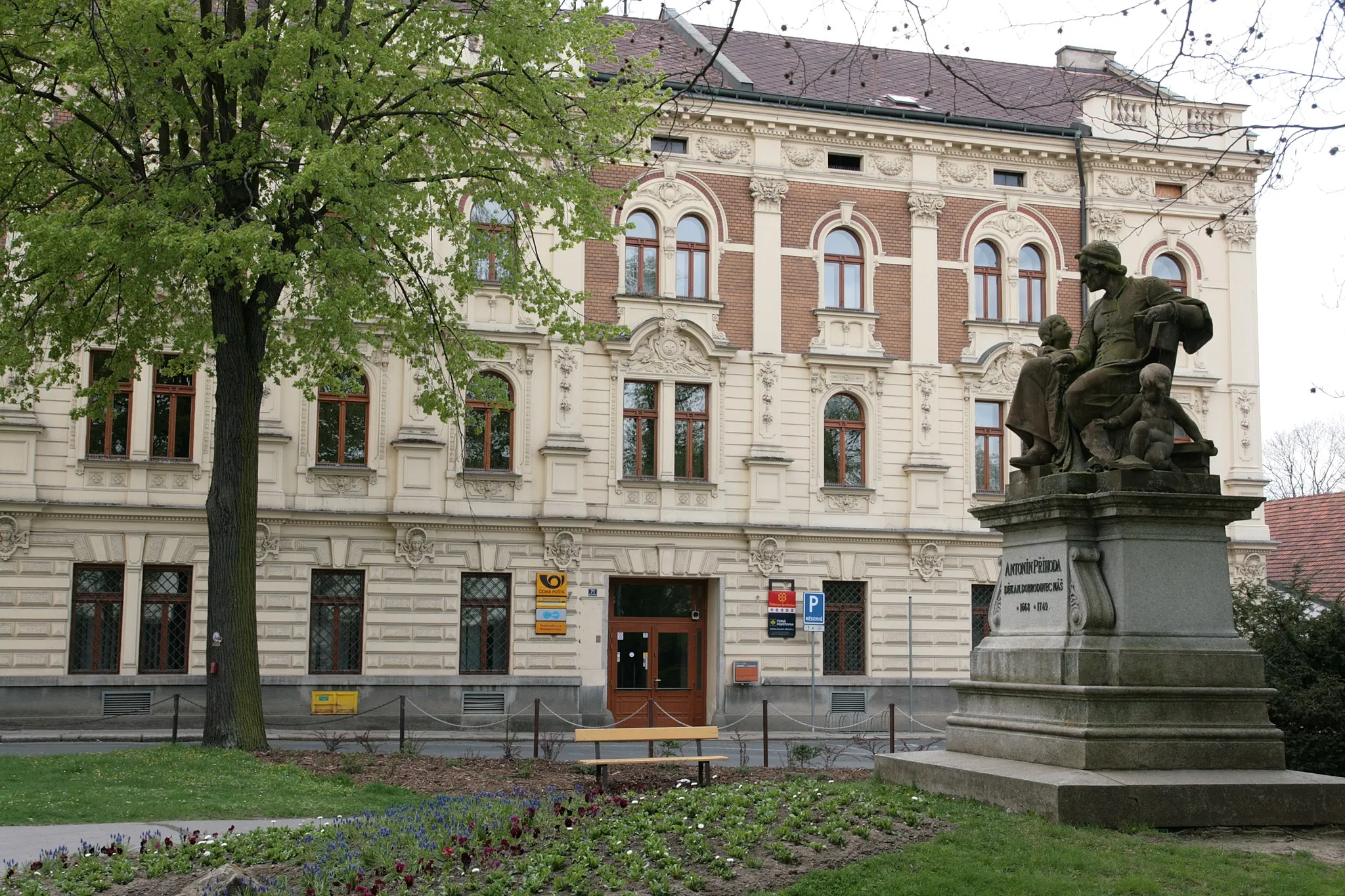 Photo showing: Město Domažlice. Pomník děkana Antonína Příhody v Příhodových sadech. V pozadí budova pošty, čp. 71 v ulici Msgre. B. Staška.