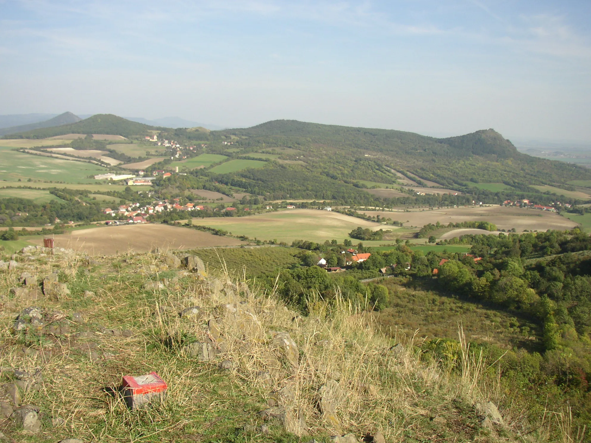 Photo showing: View from summit of Plešivec (477 m) in České středohoří Mts. towards east. In the foreground hamlet of Chrastná with wide valley of the Modla stream beyond. In the valley on the left village of Vlastislav with castle tower and chateau visible. On the right side there lies village of Teplá. Hills on the horizon left to right: Boreč (446 m), Sutomský vrch (505 m),  at centre Jezerka (487 m) and Košťál (aka Košťálov, 481 m) with another castle ruin.