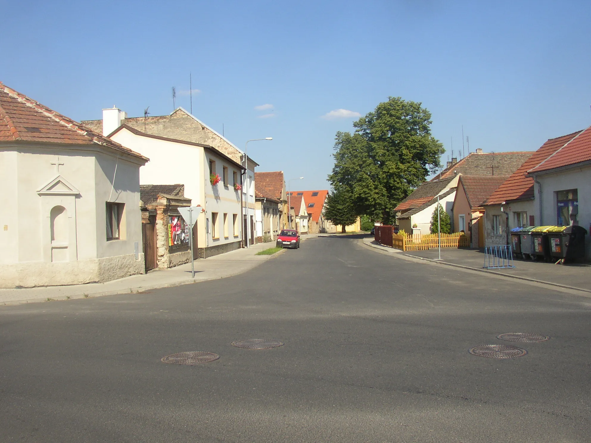 Photo showing: Brňany, Litoměřice District. A view from crossroads of main streets towards east. In the left small niche chapel in corner of house No 30, in the background WWI memorial hidden in shade of big trees.