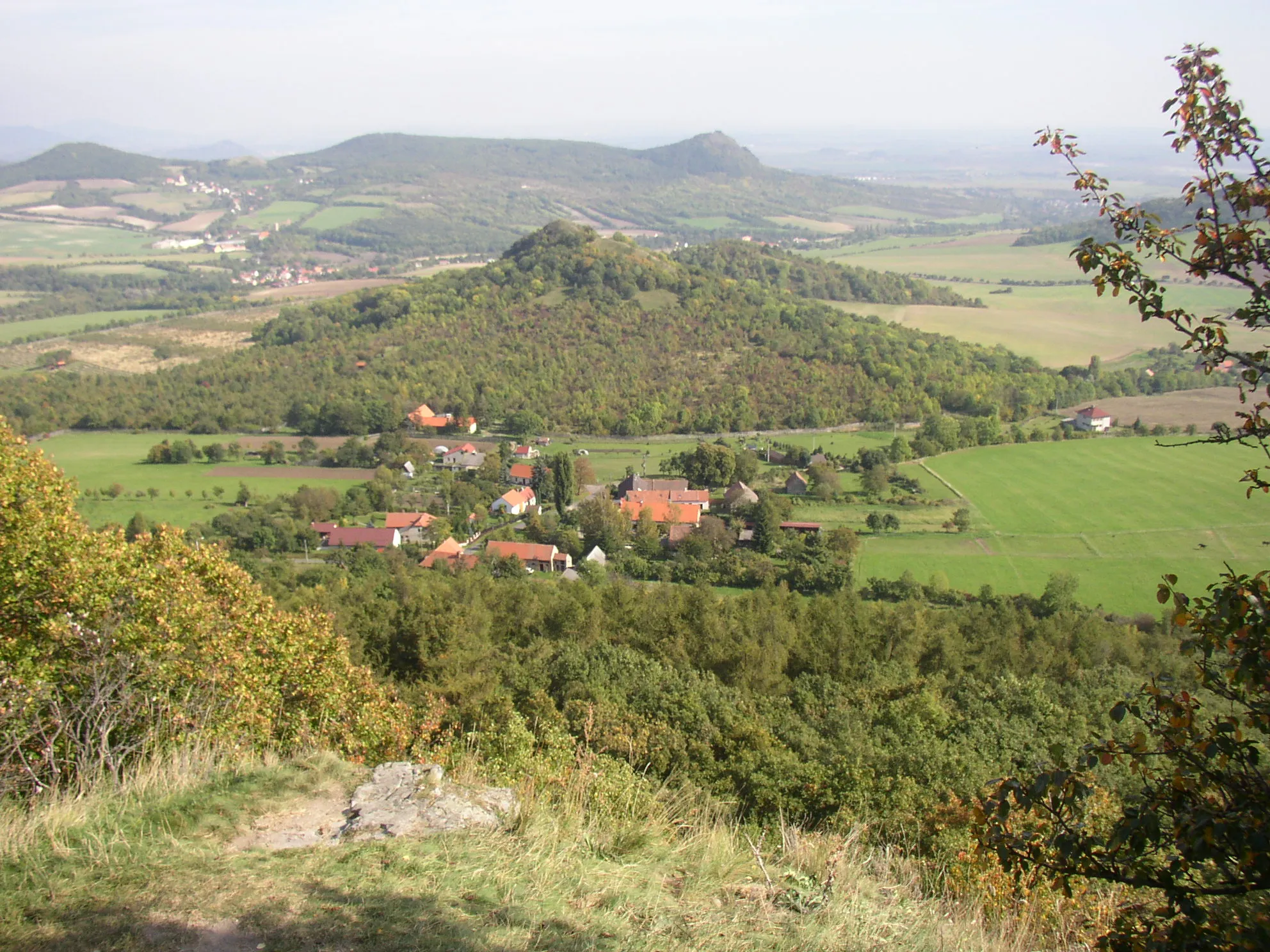 Photo showing: View from ruins of Oltařík Castle in České středohoří Mts. towards east. In the foreground village of Děkovka with hill of Plešivec (477 m) beyond it. On the horizon slightly right off the centre there is hill of Košťál (aka Košťálov, 481 m) with another castle ruin and in the left hill of Sutomský vrch (505 m) with villages of Sutom on its slopes and Vlastislav at foothill..