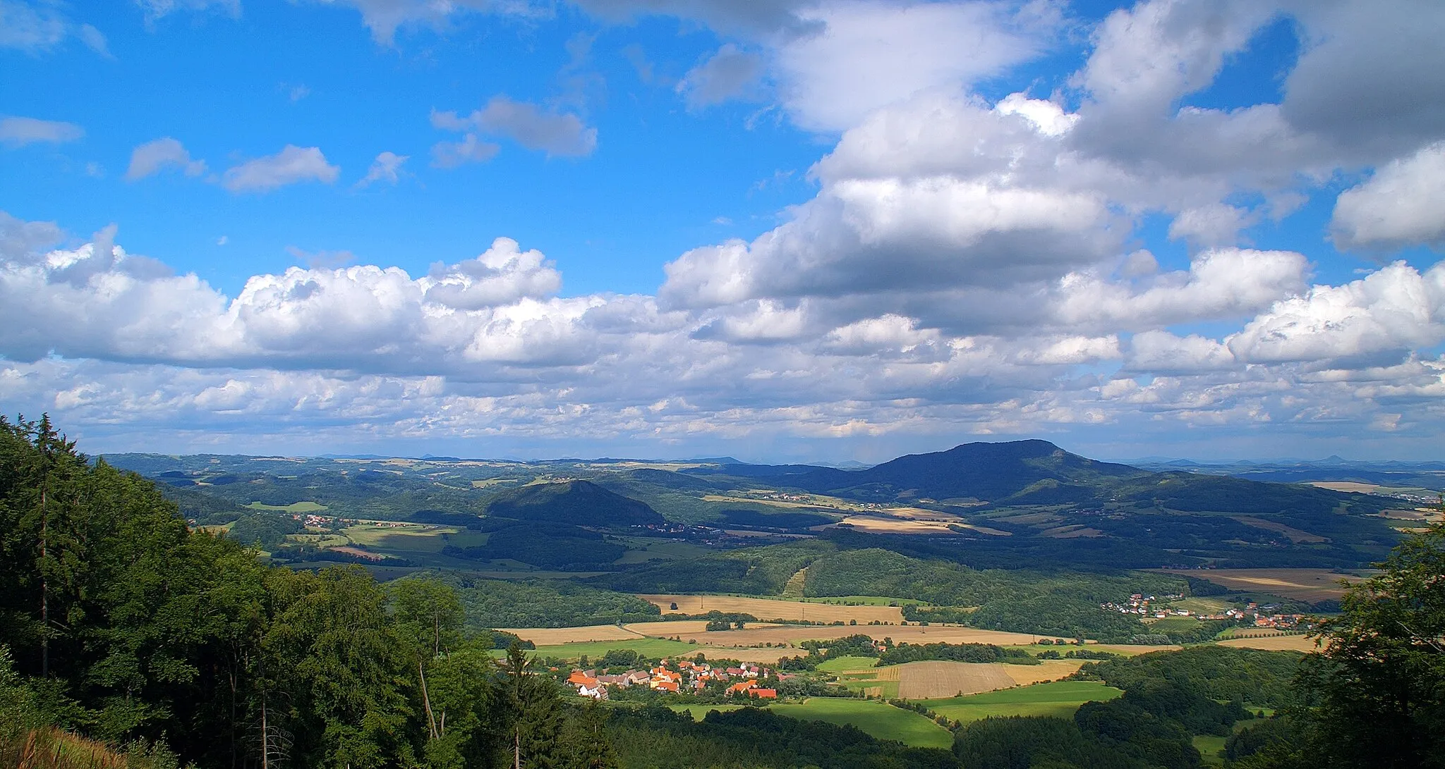 Photo showing: Sedlo (v pravé polovině snímku) je nejvyšším vrchem východní části Českého středohoří (726 m.n.m.). Snímek je pořízen z hřebene Dlouhého vrchu. Kotlina mezi oběma horami je významnou ovocnářskou oblastí a někdy je proto nazývána českou Kalifornií. Dole uprostřed snímku je obec Staňkovice.