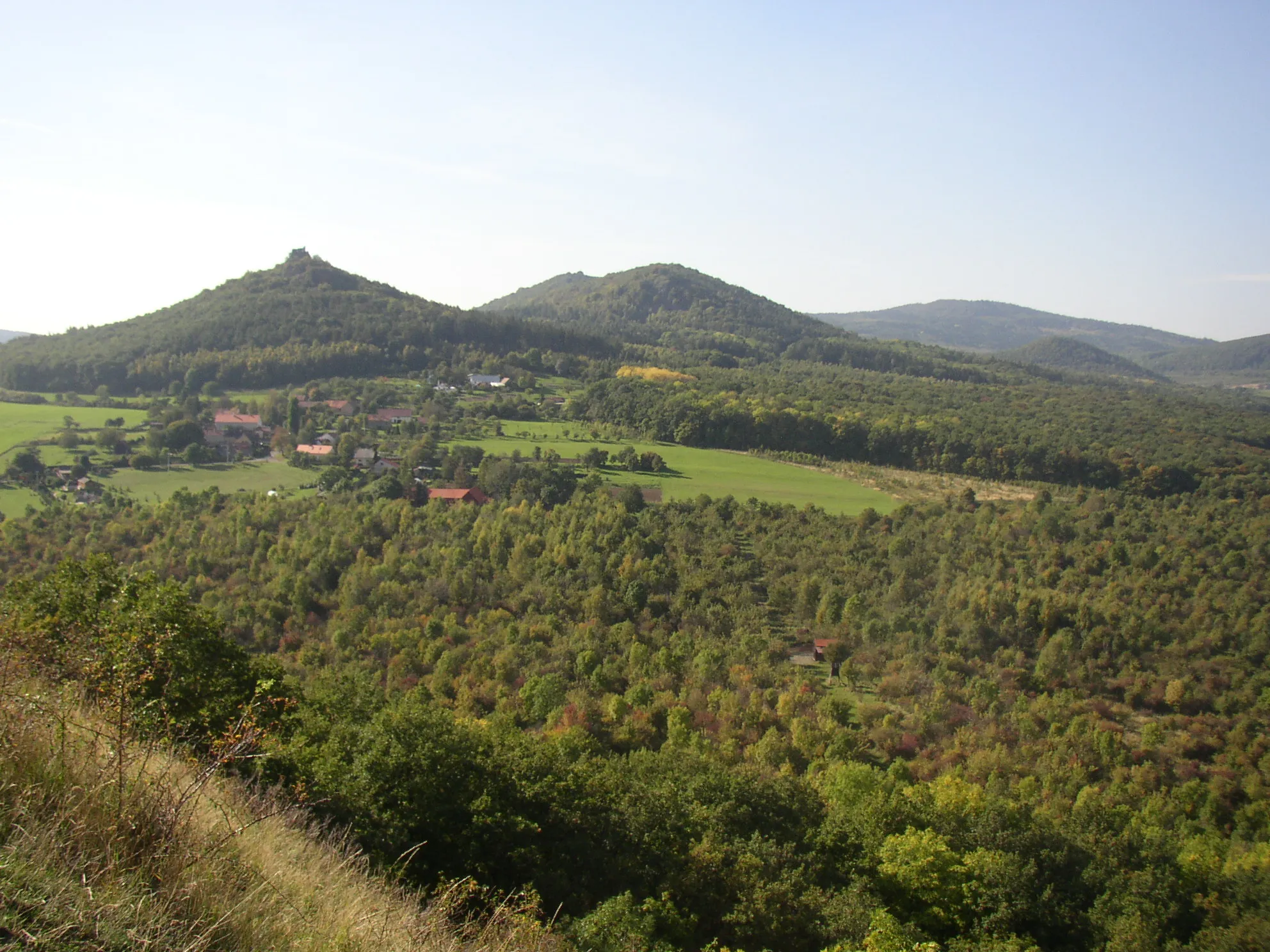 Photo showing: View from summit of Plešivec (477 m) in České středohoří Mts. towards west-northwest. In the left above village of Děkovka rises hill of Oltářík (565 m) with castle ruin on its top. The elongated double summit in the middle belongs to Solanská hora (638 m), the right horizon is filled with huge shape of Hradišťany (752 m). At its foothill there appears much smaller hill of Líšeň (548 m).