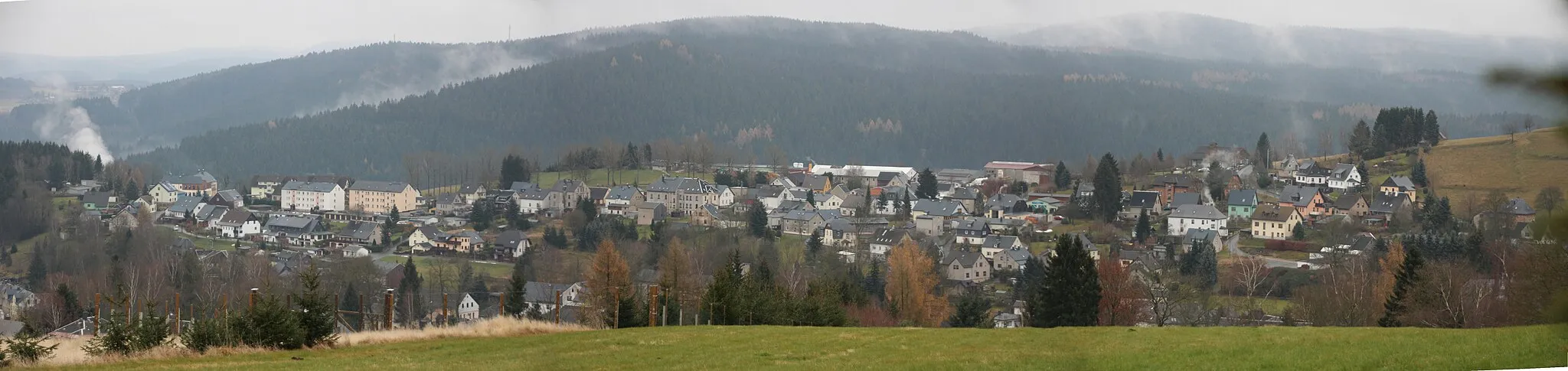 Photo showing: Schönheide im Erzgebirge: Ortsteil Schädlichsberg von der baumlosen Erhebung in der Ortsmitte Allee, früher auch Oberförsterknock genannt (684 Meter), aus. Der Schädlichsberg liegt auf einem plateauartigen, nach Osten abfallenden  Sporn mit West-Ost-Verlauf  zwischen dem Dorfbach, an dem sich das Dorf in seinem langgestreckten Hauptteil hinzieht, und der Zwickauer Mulde. Dieser Sporn ist ein Ausläufer des Berges Knock (725 Meter), der südlich des mittleren Ortsteil liegt. Hinter den Häusern des Ortsteils Schädlichsberg ist deutlich der tiefe Einschnitt des Tals der Zwickauer Mulde zu sehen. Der (linke) östliche Teil des Schädlichsbergs trug früher die Ortsbezeichnung Buchhöhe (erzgebirgisch Guchheh). Er gehörte zum heutigen Schönheider Ortsteil Schönheiderhammer. Etwa in der Mitte des Ortsteils wird das Uttmannsche Vorwerk, das zu Schönheiderhammer gehörte, gelegen haben. Sein indirekter Nachfolger war in der DDR-Zeit die LPG Vorwärts, sie hatte ihre Rinderstallungen, Scheunen und anderen Baulichkeiten in der Mitte des Fotos (flache hellgraue Dächer, z. T. Nachfolgebauten). Rechts am Rand der Berg Knock, im Hintergrund die Berge jenseits der Zwickauer Mulde. Fotografiert vom Berg Allee aus.