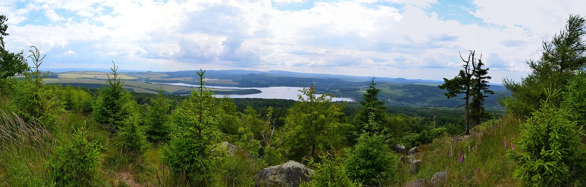 Photo showing: Panoramablick vom Gipfelrand des Jelení hora von Süden nach Nordwest. Im Hintergrund ist das Fichtelberg-Keilberg-Massiv, davor der Velký Špičák und im Vordergrund Stausee sowie Damm der Talsperre Preßnitz erkennbar. Unterhalb des Dammes liegt die Ortschaft Kryštofovy Hamry. Im Süden lässt sich zudem der Mědník sowie der Steilabbruch des Gebirges zum Egergraben hin erkennen.