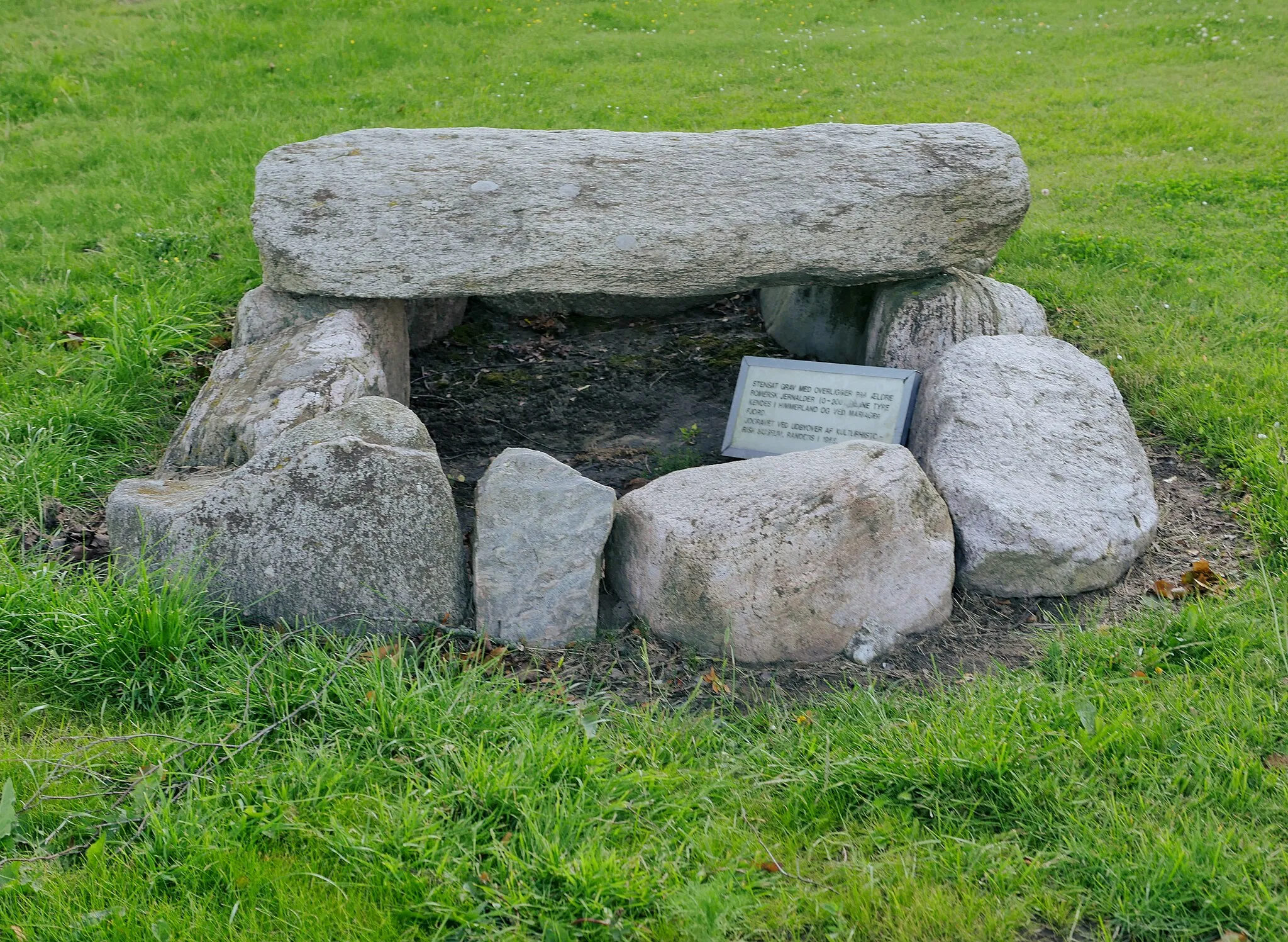 Photo showing: Havndal Steinkistengräber Grab 3
Eine weitere Steinkiste wurde östlich von Udbyover gefunden. Sie liegt wiederaufgebaut im Havndal Sportzentrum. Dieses aus Findlingen errichtete Grab ist ebenfalls Ost-West-orientiert. Über der Mitte lag eine große, flache, durch den Pflug verkratzte Platte. Die Kiste misst innen 2,1 × 1,4 m, die Tiefe beträgt 0,5 m. Diese Art von Steinkiste ist eine am Mariagerfjord übliche Mischung aus der großen Steinkiste Nord-Jütlands und den grazilen Plattenkisten in Ost- und Mitteljütland. Die Mariager Steinkisten sind nicht so massiv wie die in Nordjütland errichteten. Die Position und der Aufbau der Tonwaren ist nicht so festgelegt wie in Ost- und Mitteljütland. Waffen sind in diesen Gräbern selten. Gefunden wurden eine 0,5 m lange Lanzenspitze aus Eisen, ein Bronzering und Scherben von 8 bis 9 Keramiken aus der frühen römische Eisenzeit. Der Tote war ohne Zweifel ein Mann. Er liegt im Nordabschnitt des Grabes, mit dem Kopf nach Westen. Spuren einer Siedlung sind in der Nähe gefunden worden.