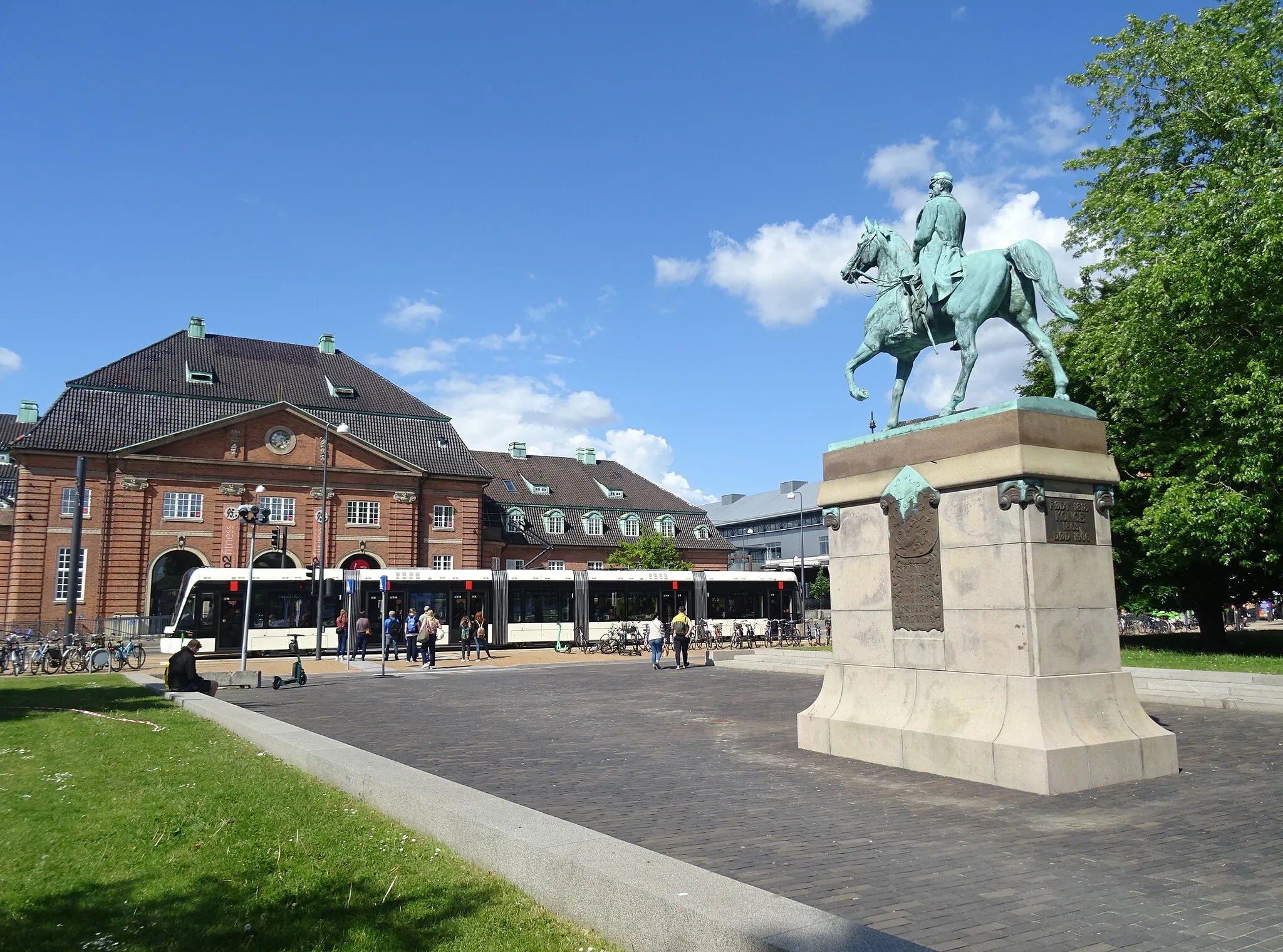 Photo showing: A Stadler Variobahn of Odense Letbane on Østre Stationsvej between the former station building and the equestrian status of king Christian IX in Odense in Denmark.