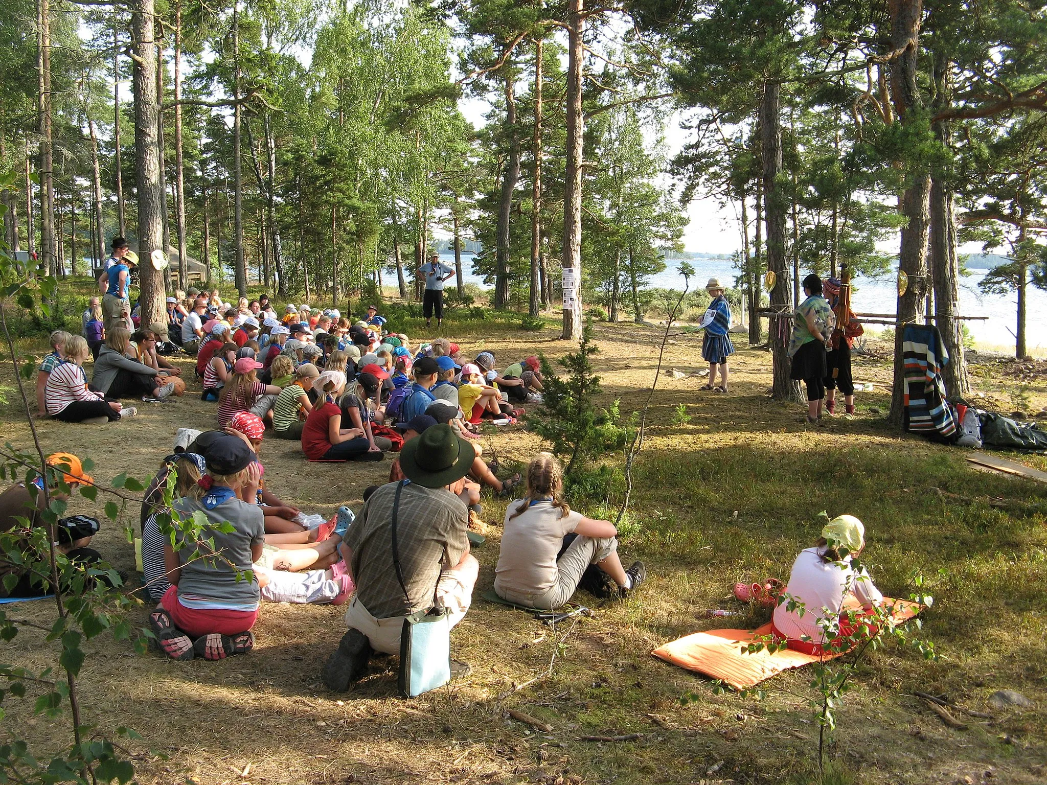 Photo showing: "Inkas" explaining how to make a piece of jewelry on the joint summer camp of the scouts of South-Western Finland 2009, "Reila". Archipelago of the Bothnian Sea in the background.