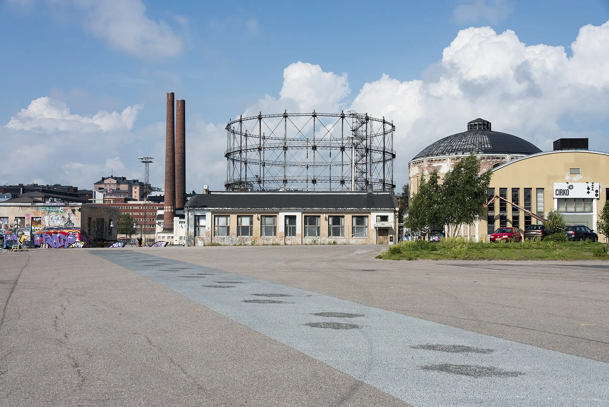 Photo showing: Kalasataman dokumentointi. Suvilahden voimalaitoksen alue nähtynä idästä uuden Kalasataman asuinalueen suunnasta, kaasukellot ja voimalaitoksen rakennuksia, vasemmalla graffitiseinä. -- digitaalinen kuva, dng ja tiff, väri