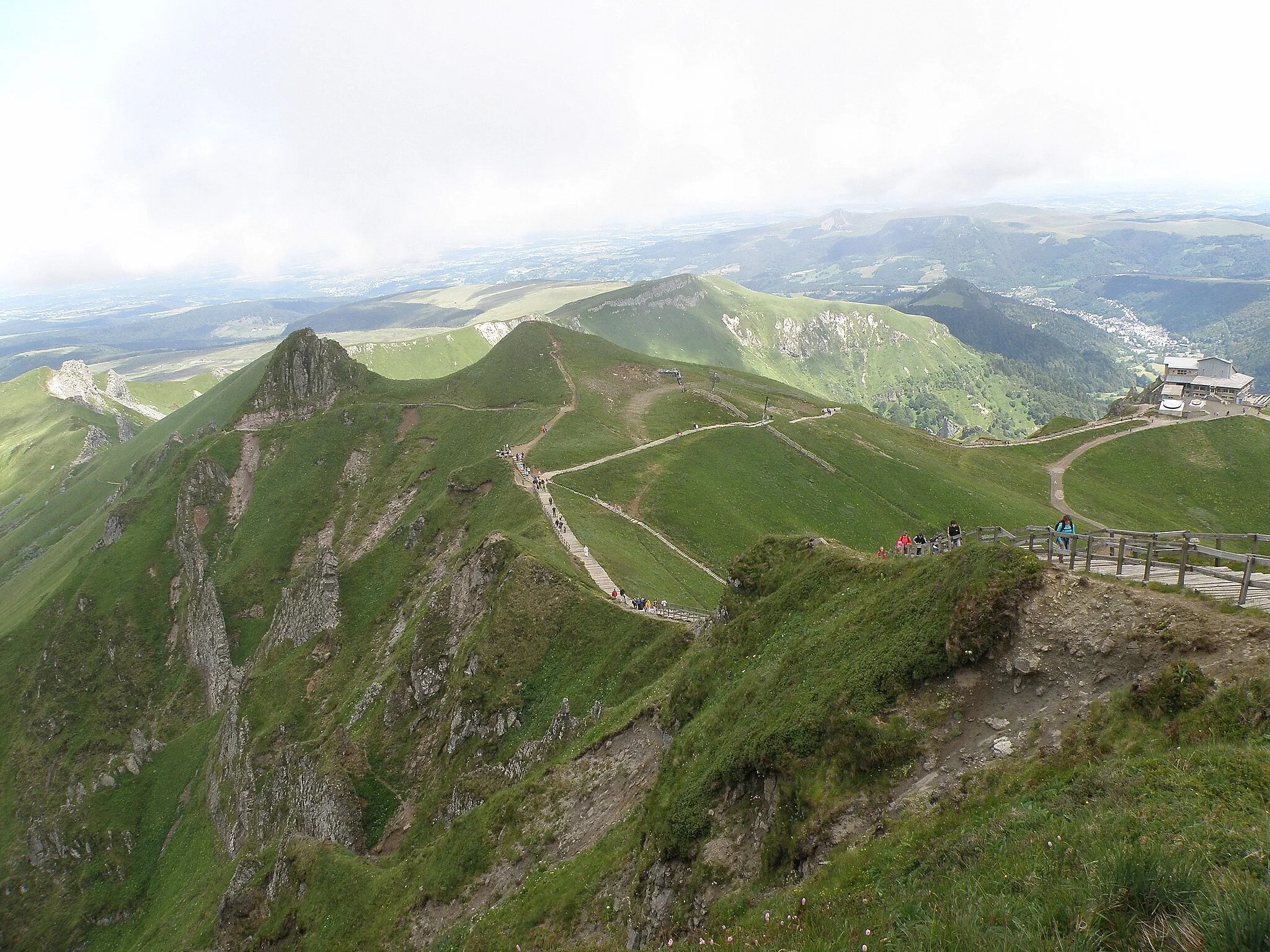 Photo showing: Le Mont-Dore, comm. du Puy-de-Dôme (France, région Auvergne). Massif du Sancy. Photographie prise depuis le flanc nord du puy de Sancy. Coup d'œil vers le nord. Chemin de madriers conduisant de la station du téléphérique, visible tout à droite, vers le sommet du puy de Sancy, lequel se trouve derrière nous. À gauche, paroi à pic délimitant le cirque de la Fontaine salée, lequel s'étend hors de la photo vers la gauche (c.-à-d. au sud-ouest). Derrière les deux pics coniques à gauche, s'aperçoivent, un peu surexposés, de gauche à droite : l'Aiguille (contre le bord gauche de la photo), les rochers de l'Hor (entre les deux pics), le puy de Cliergue (au centre), et le sommet arrondi du Capucin (dans l'ombre d'un nuage). Dans le fond à droite, derrière la station du téléphérique, ville du Mont-Dore.