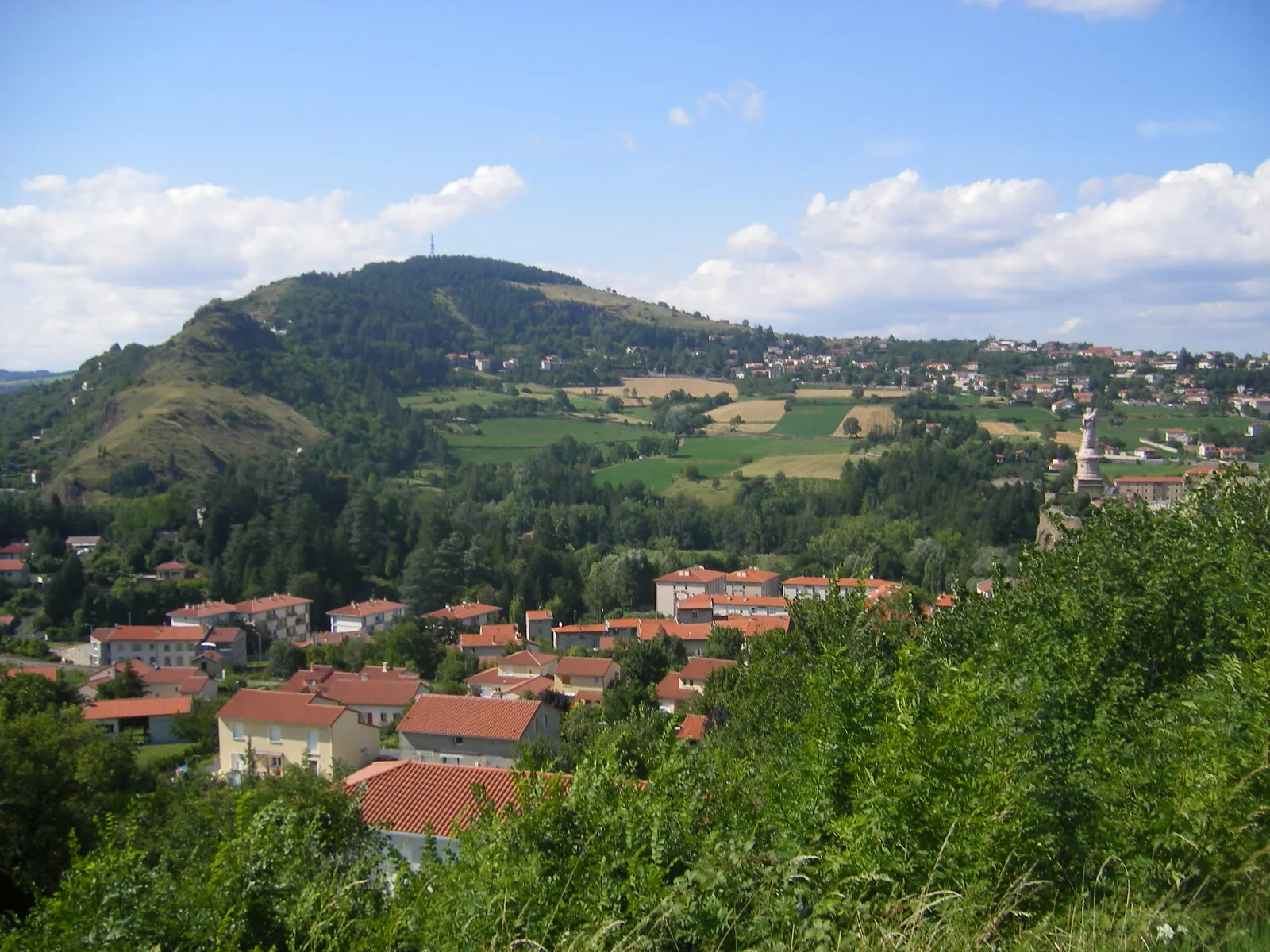 Photo showing: Espaly-Saint-Marcel (Haute-Loire, south Auvergne, France). Looking north-west from the D589 road. On the right-hand edge are the statue and basilica of the Saint-Joseph-of-Good-Hope (Saint-Joseph-de-Bon-Espoir) sanctuary (which also includes a chapel in a grotto). In the background, the inactive volcano of mont Denise with it antenna at the top (alt. 892 m). The mount on the left is the "Orgues d'Espaly" (alt. 783 m).