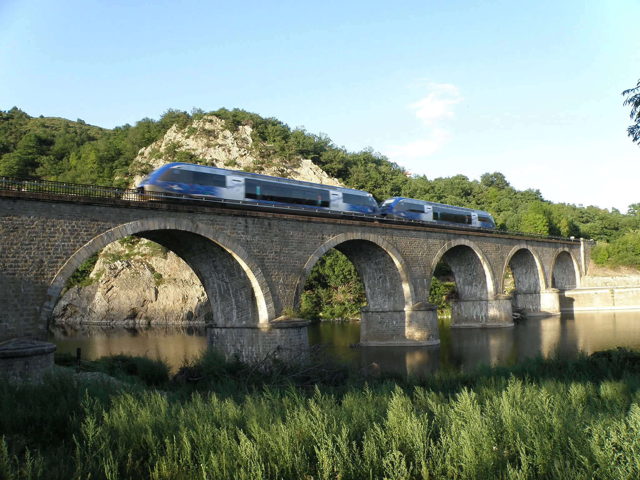 Photo showing: Beauzac, dép. de la Haute-Loire, France (Auvergne). Viaduc ferroviaire de Brenas, près du hameau de Brenas (dont on aperçoit la toiture d'une des maisons derrières les cimes des arbres au centre droit), dans le sud de la commune. Un train de type TER (Train Express Régional) faisant le service entre le Puy et Saint-Étienne franchit la Loire, qui fait ici frontière avec la commune voisine de Retournac. Les falaises à l'arrière-plan à gauche sont les Côtes du Suc.