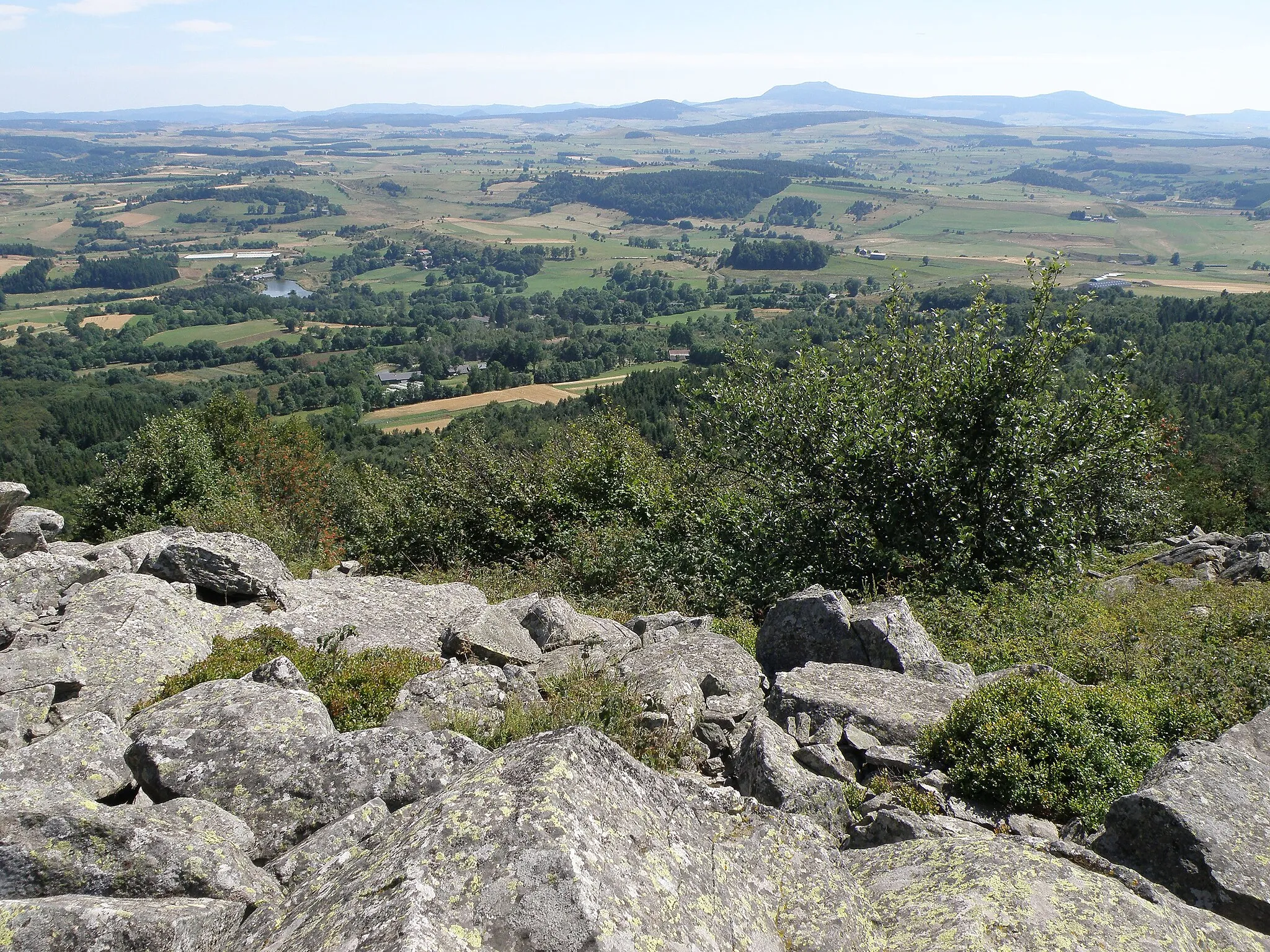 Photo showing: Araules, commune de la Haute-Loire, France, région Auvergne. Vue prise depuis le pic du Lizieux (appelé aussi suc du Lizieux, alt. 1388 mètres, rochers à l’avant-plan), sis dans le sud-est de la commune d’Araules. La plaine en contrebas, du moins sa partie plus rapprochée, appartient à la comm. de Mazet-Saint-Voy; seule une petite étendue contre le bord droit de la photo se situe dans la comm. d’Araules. L’étang visible dans la plaine à gauche se trouve à l’emplacement d’une ancienne mine ; le groupe de maisons un peu à droite de l’étang est le hameau du Bouchat. Un peu plus près de nous : le hameau de Faurie (toitures grises).
À l’horizon, le sommet le plus élevé sur la photo (et dans la réalité) est le mont Mézenc (alt. 1753m), reconnaissable à sa silhouette caractéristique ; à sa droite, le mont d’Alambre (alt. 1691). Contre le bord droit, tombant en partie en dehors de l’image : (probablement) le Rocher Tourte (alt. 1535). Le morne aplati que l’on aperçoit au centre, à gauche du Mézenc, et plus proche de nous, pourrait être le mont Signon (alt. 1455m, comm. de Chaudeyrolles).
Objectif orienté au sud.