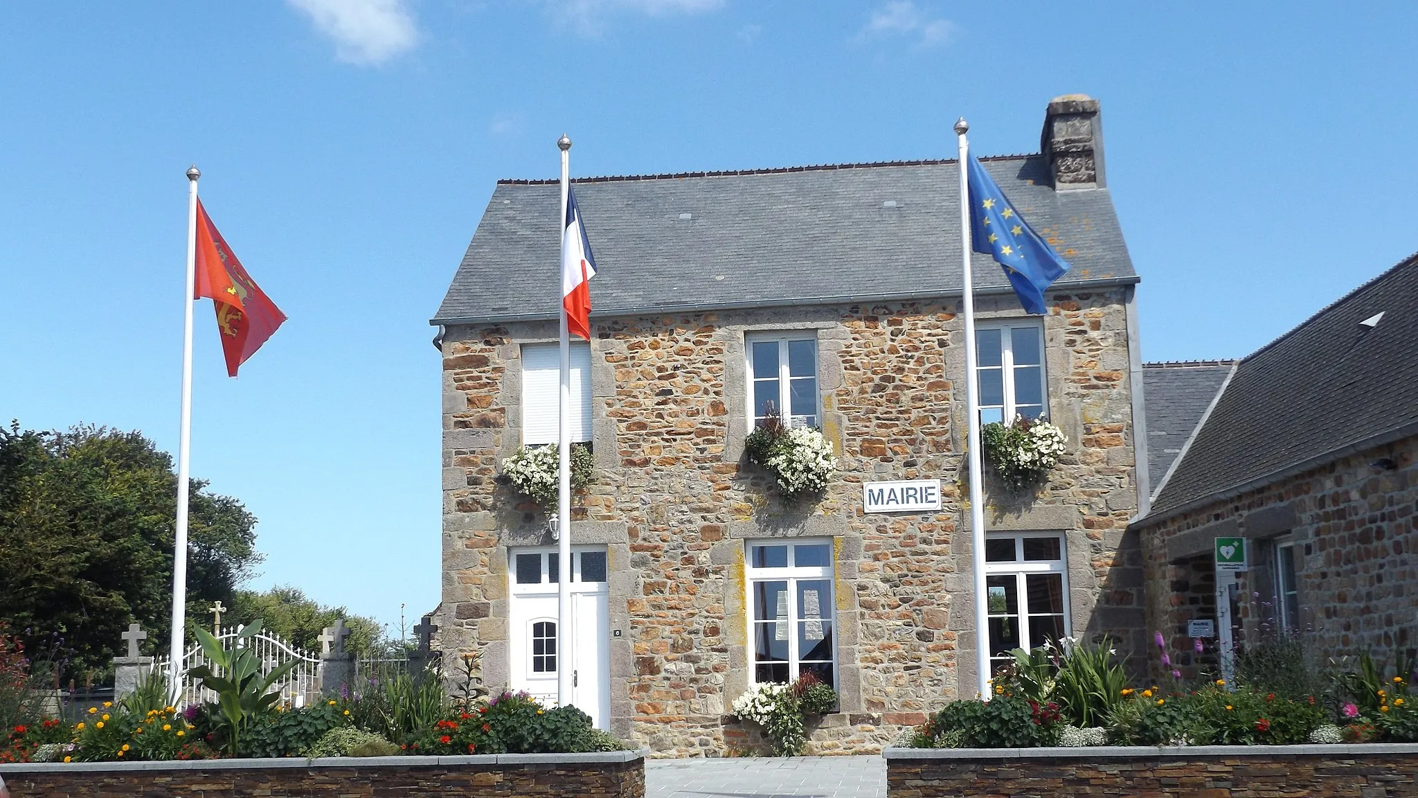 Photo showing: En premier plan les murets surmontés de fleurs, dans lesquels sont plantés les drapeaux de la Normandie, de la France et de l'Europe Les murets délimitent la parvis piéton de la mairie. En fond de champ, le bâtiment en pierre de la marie, couvert en pierre du pays, au fons à gauche, l'allée du cimetière