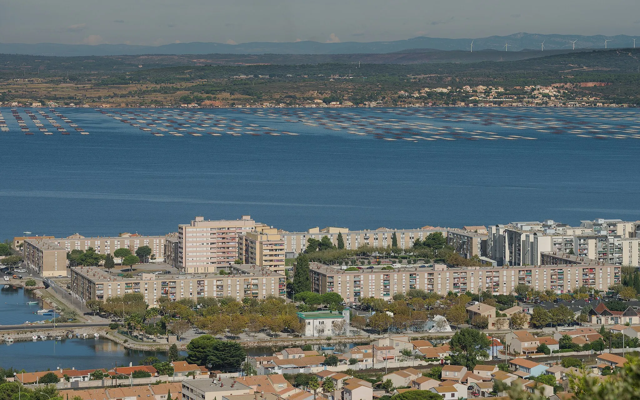 Photo showing: In the foreground the Southwestern part of the Ile de Thau Neighbourhood and the Étang de Thau.  In background on the opposite bank the commune of Loupian and its oyster farms. Sète, Hérault, France.