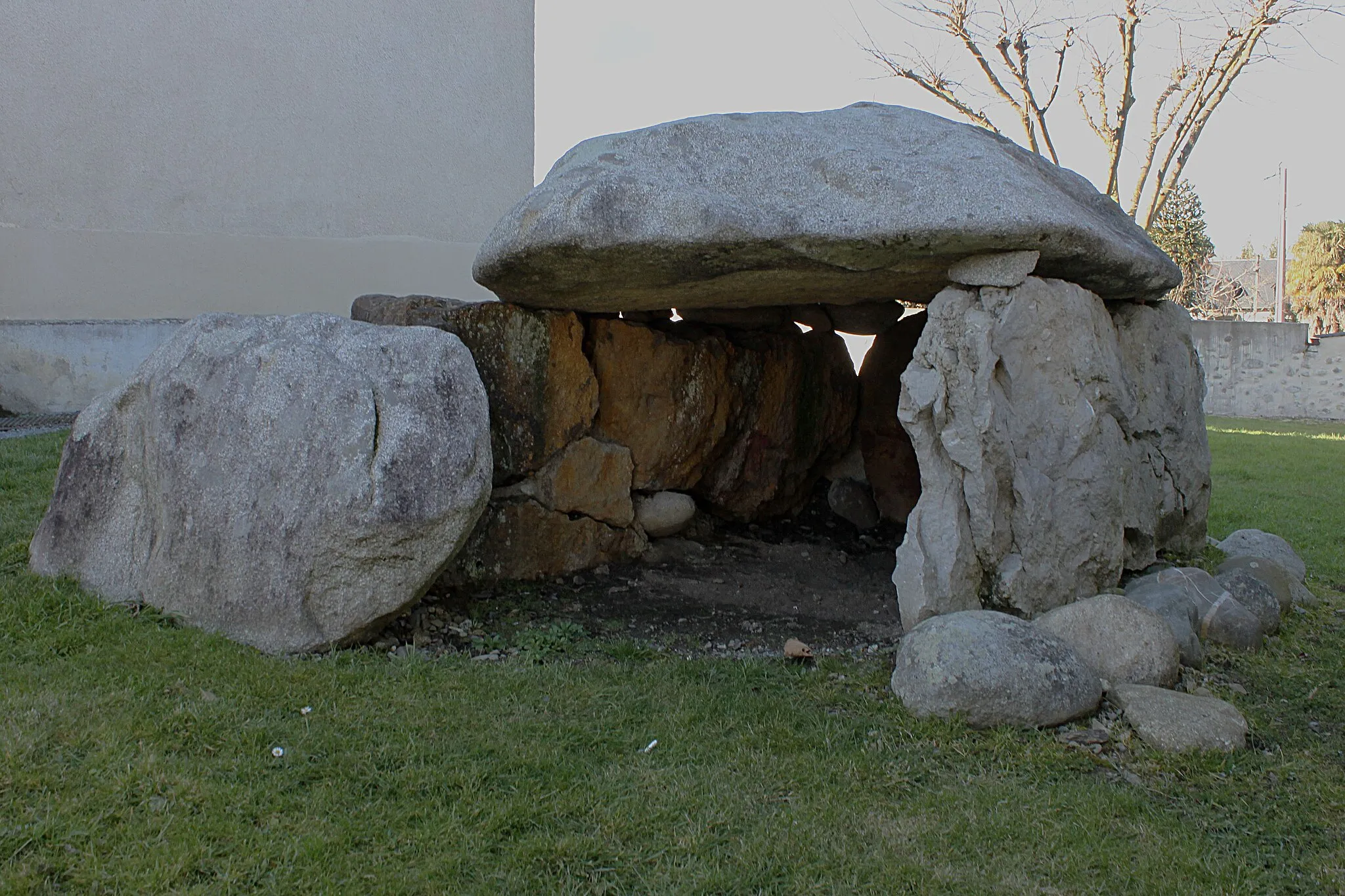 Photo showing: The dolmen of Barzun dates from 3500 to 4000 years ago, around the end of the Neolithic and start of the Bronze Age. It was discovered in 1968 in a field in Barzun. The original structure was about 20 x 10 metres
This oval burial mound had a height of 1,5 m at its highest point. As it was excavated, two tangent circles made of boulders appeared. In the second circle was discovered the dolmen itself. The room was to have received one or more burials accompanied by furniture and various valued items such as vases, polished axes, and flints. Unfortunately, the interior of the dolmen had certainly been violated at an early date, and archaeologists discovered nothing in it, neither bones nor vases or worked stone.
The dolmen was then dismantled in 1968 and moved in 1972 to Arudy (a village about 50 km from Barzun) by Georges Laplace, research fellow at the CNRS. In 1990, Laplace moved from Arudy to Coarraze and gave the dolmen to the Municipality of the commune, which installed it on the Town Hall Square.
Barzun, in 1995, tried to have the dolmen brought back to the village, but with no success.
In 2010, a understanding was reached between Jean Saint-Josse (the Mayor of Coarraze) and , Maurice Minvielle (Mayor of Barzun). Coarraze Council then authorised the return of the dolmen to its barzunaise origins.
It was described in several stories as "the travelling dolmen."

The transfer from Coarraze took place in January 2011, and the installation on an old cemetery by the church of Barzun took place on January 13, 2012.