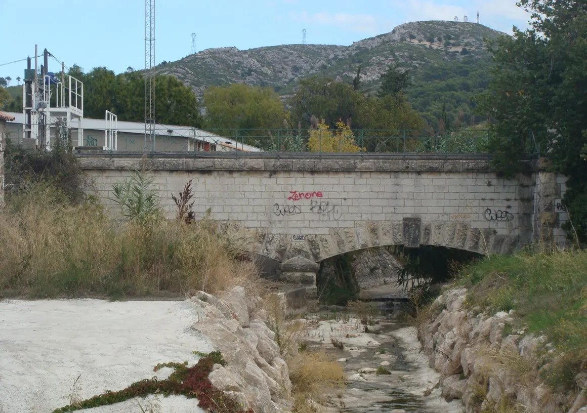 Photo showing: Petit aqueduc à 2 arches en pierre par lequel le canal de Marseille franchit le Jarret à la limite entre Allauch et Plan-de-Cuques. Vue au zoom vers l'est depuis la rue de l'Hermitage. A gauche derrière le canal, une partie des installations de l'espace culturel et sportif Robert Ollive. En arrière-plan, les hauteurs de la Tête Blanche où passent deux lignes d'électricité à très haute tension.