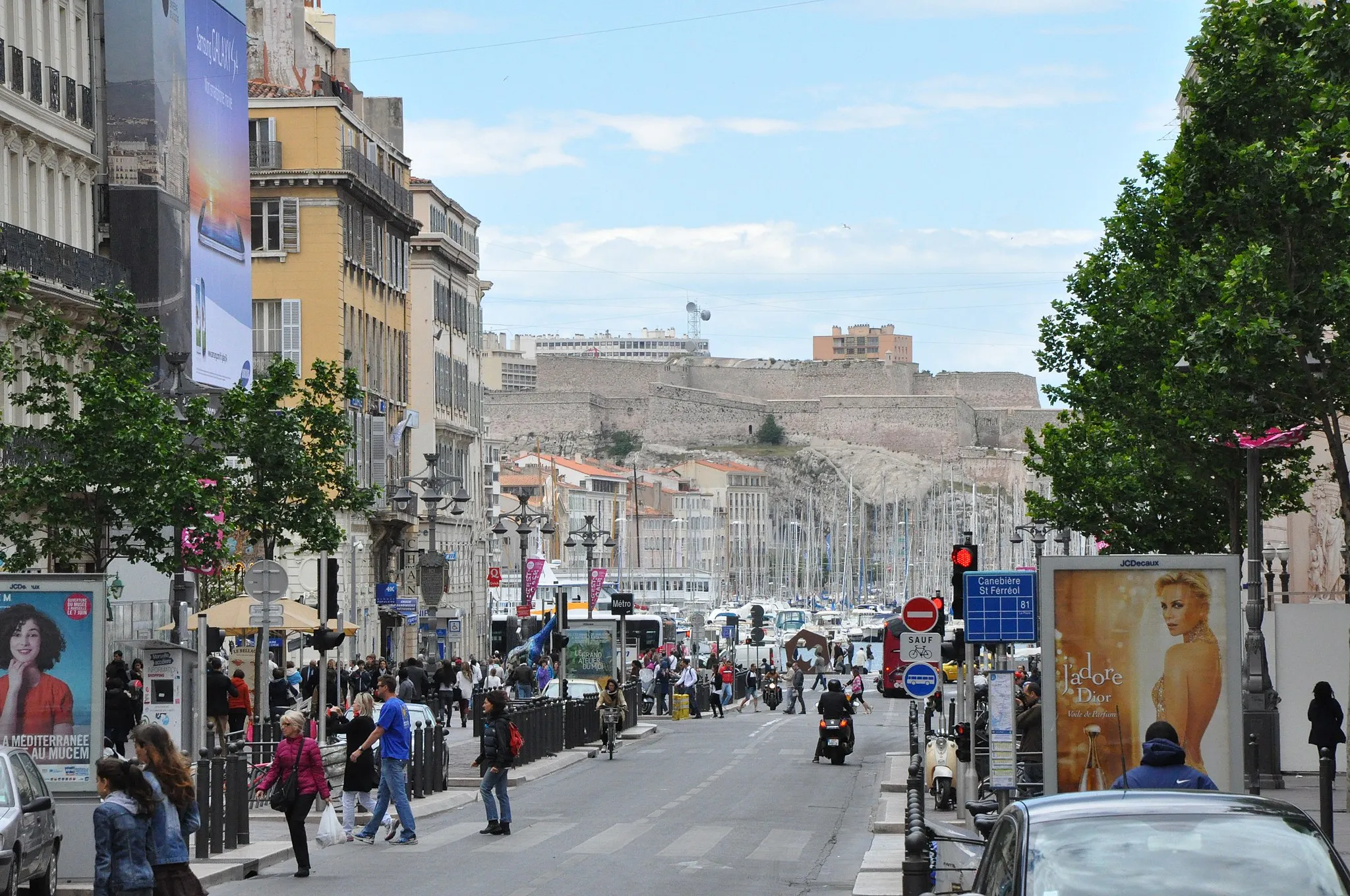 Photo showing: La Canebière, between quarters Opéra (left part of the image) and Hôtel-de-Ville (right part of image) in the 1st Arrondissement, Marseille, Provence, France. Seen from the intersection with Cours Saint-Louis (to the right) and Cours Belsunce (to the left), in direction of Vieux-Port, then Fort Ganteaume which borders it to the south-west. In the background, the Saint-Nicolas Fort (in quarter of Pharo) dominates the west of the Vieux-Port and to the south-east the quarter of Saint-Victor (unvisible, to the left of this image).