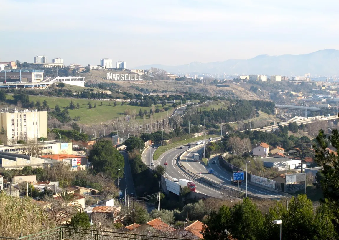 Photo showing: Le versant ouest de la dorsale de la Viste à Marseille (16e arrondissement), avec à gauche le centre commercial Grand Littoral. Derrière au centre, l'inscription « MARSEILLE » en lettres géantes posées dans le parc Foresta, au flanc du Belvédère de la Viste. Sur la gauche, quelques bâtiments du bas du quartier de la Pelouque et en bas de l'image un tunnel sous l'autoroute relie les deux parties du quartier. Vu des hauteurs du hameau de la Pelouque séparé par l'autoroute A55 qui contourne le reste du quartier de Saint-Henri (15e arrondissement). La sortie tournant vers la droite descend vers le quartier Saint-André (non visible sur l'image).