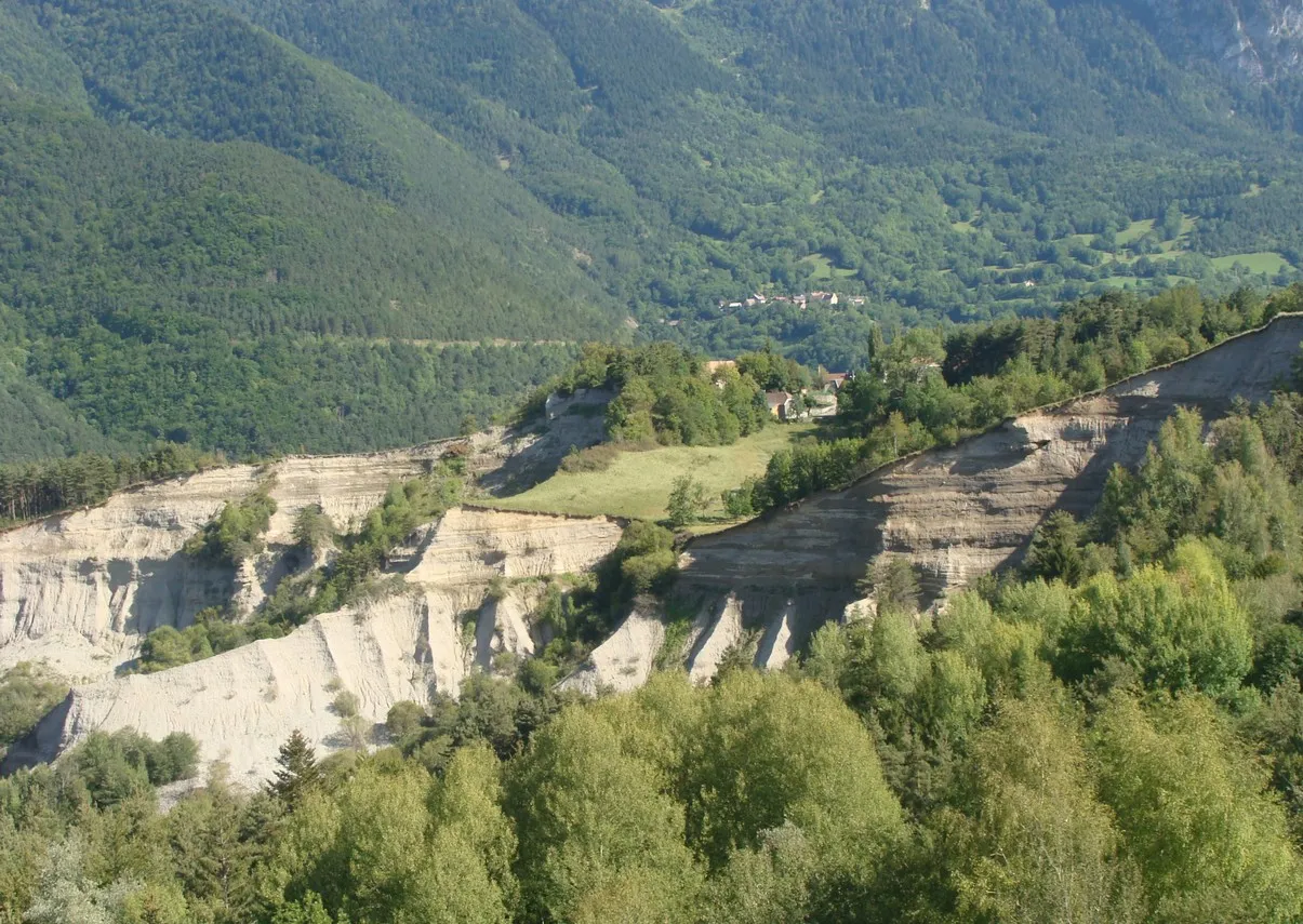 Photo showing: Le bord du plateau alluvial de Pellafol (Isère) érodé par la Souloise coulant en contre-bas. Dans les arbres au centre on aperçoit le hameau de Vieux-Pellafol, et au loin le Monestier-d'Ambel sur l'autre rive.