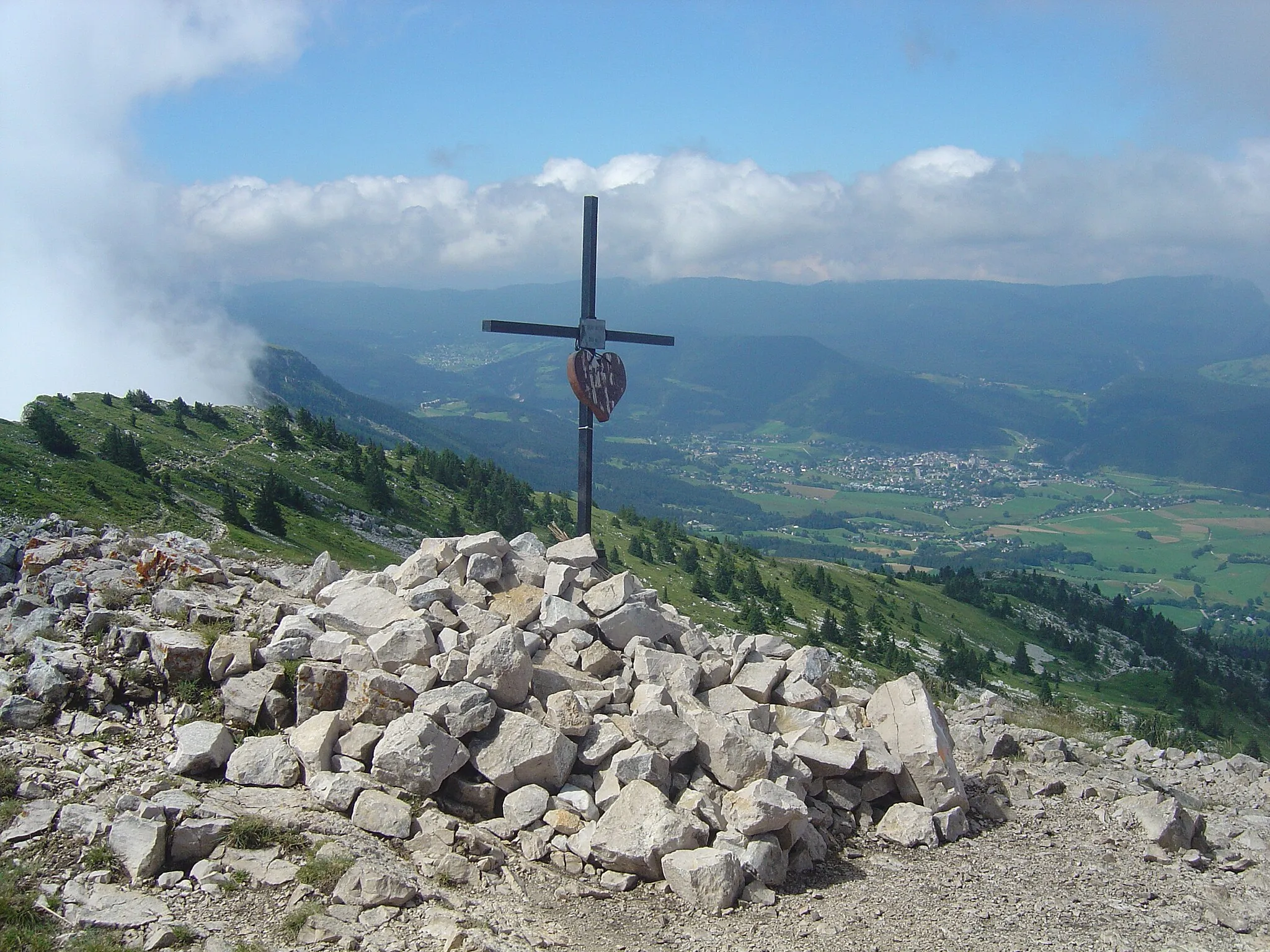 Photo showing: In the foreground on a hilltop there is a cross on a pile of stone. In the background there is a village in a valley, and in the distance there are mountains