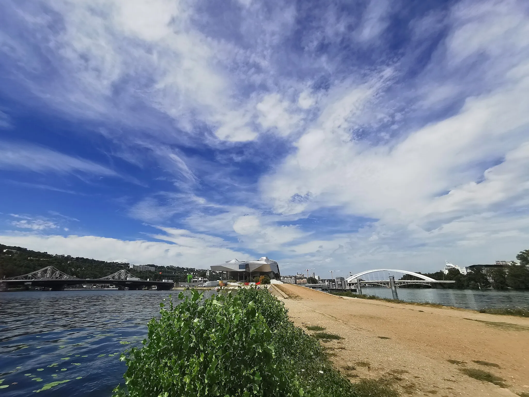 Photo showing: Photo taken at the farthest point of the Lyon peninsula (at the confluence of the Rhone and the Saône).  On the left, the Saône and the Mulatière bridges.  In the center, the Confluences museum. On the right, the Rhône and the Raymond Barre bridge.  Farthest to the right, the Docteur Charles Mérieux Lycée (which opened in September 2021). The green Euronews building is just visible on the right of the museum, and the Fourvière Basilica is just visible to its left (in the distance).