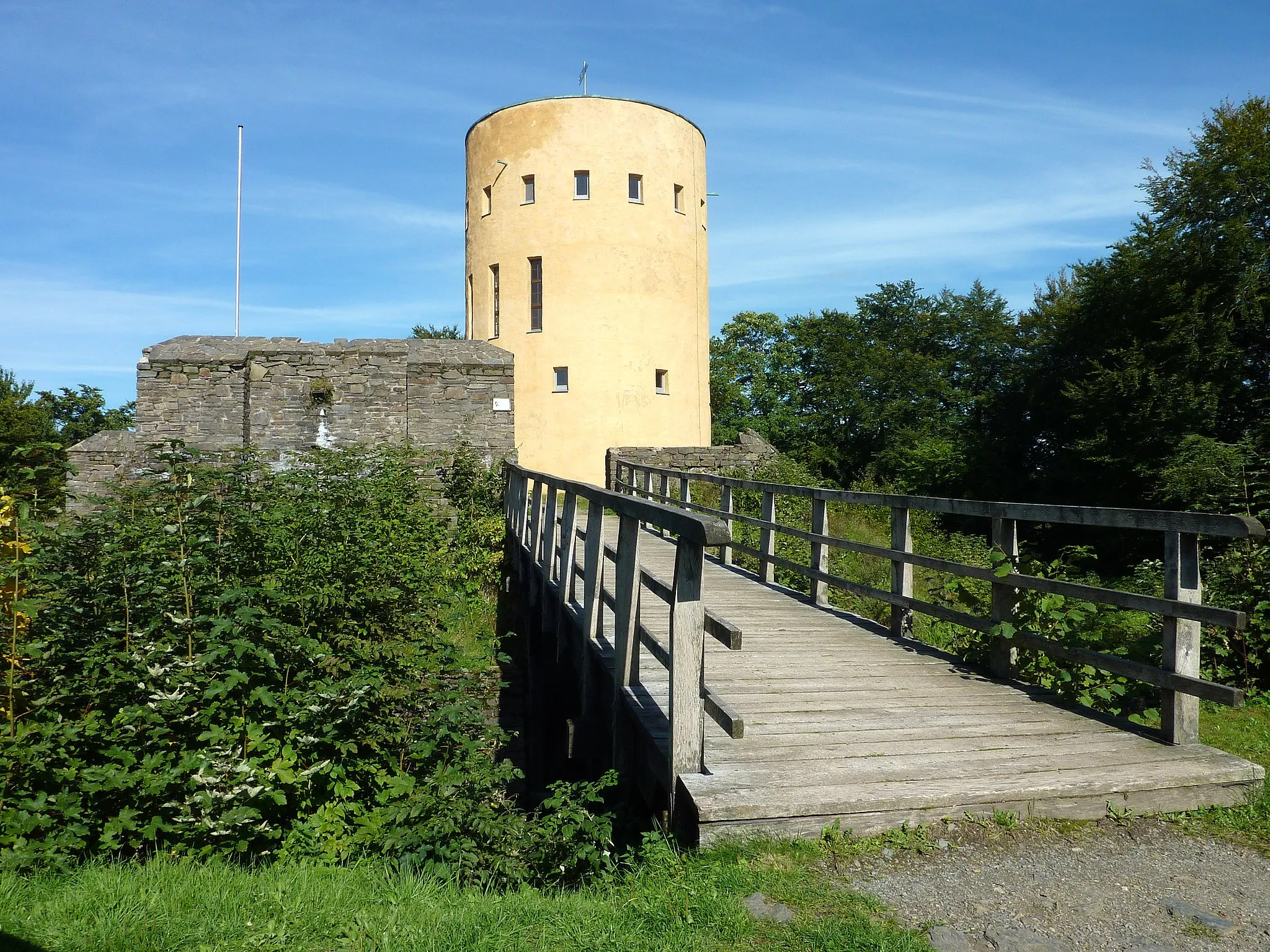 Photo showing: Gemeinde Grund (Hilchenbach)/Westfalen: Die Ginsburg-Ruine, vom 13. bis 17. Jahrhundert Grenzfestung des Hauses Nassau auf dem Schlossberg, Ansicht von Südwesten. Links im Bild ein Teil des rekonstruierten äußeren Mauerrings. In der Bildmitte der in den Jahren 1967/68 erbaute Aussichtsturm, den ehemaligen Bergfried nachahmend und auf dessen etwa aus dem Jahr 1200 stammenden Fundamenten errichtet. Im Vordergrund eine in der Gegenwart errichtete Holzbrücke über den Burggraben.