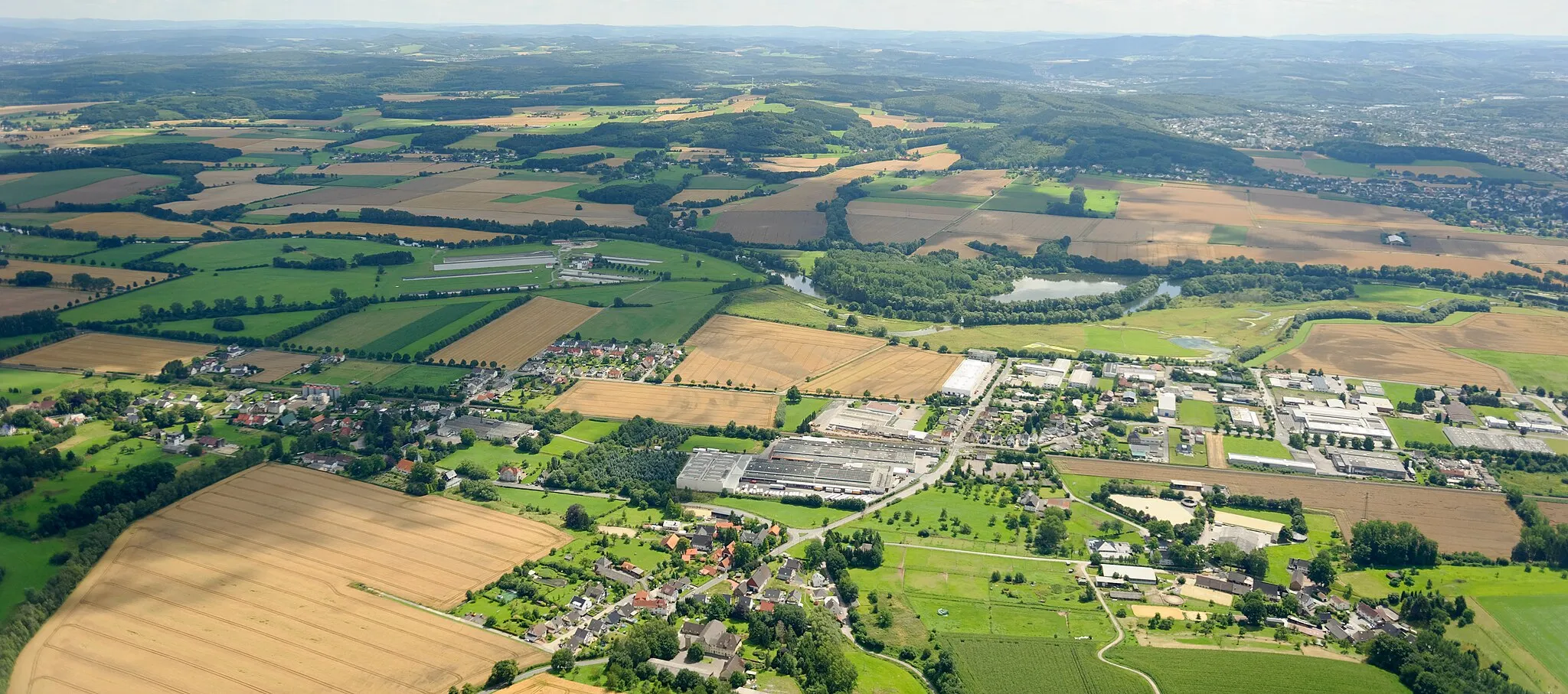 Photo showing: Luftaufnahme: Frohnhausen mit dem Industriegebiet von Fröndenberg, weiter links Warmen (mit NSG „Wulmke“), im Hintergrund die Ruhr mit NSG „Kiebitzwiese“ und dem Ententeich (NSG „Auf dem Stein“) sowie Wassergewinnungsanlage bei Warmen, Nordrhein-Westfalen. Vordergrund im Stadtgebiet von Fröndenberg, Hintergrund im Stadtgebiet von Menden.