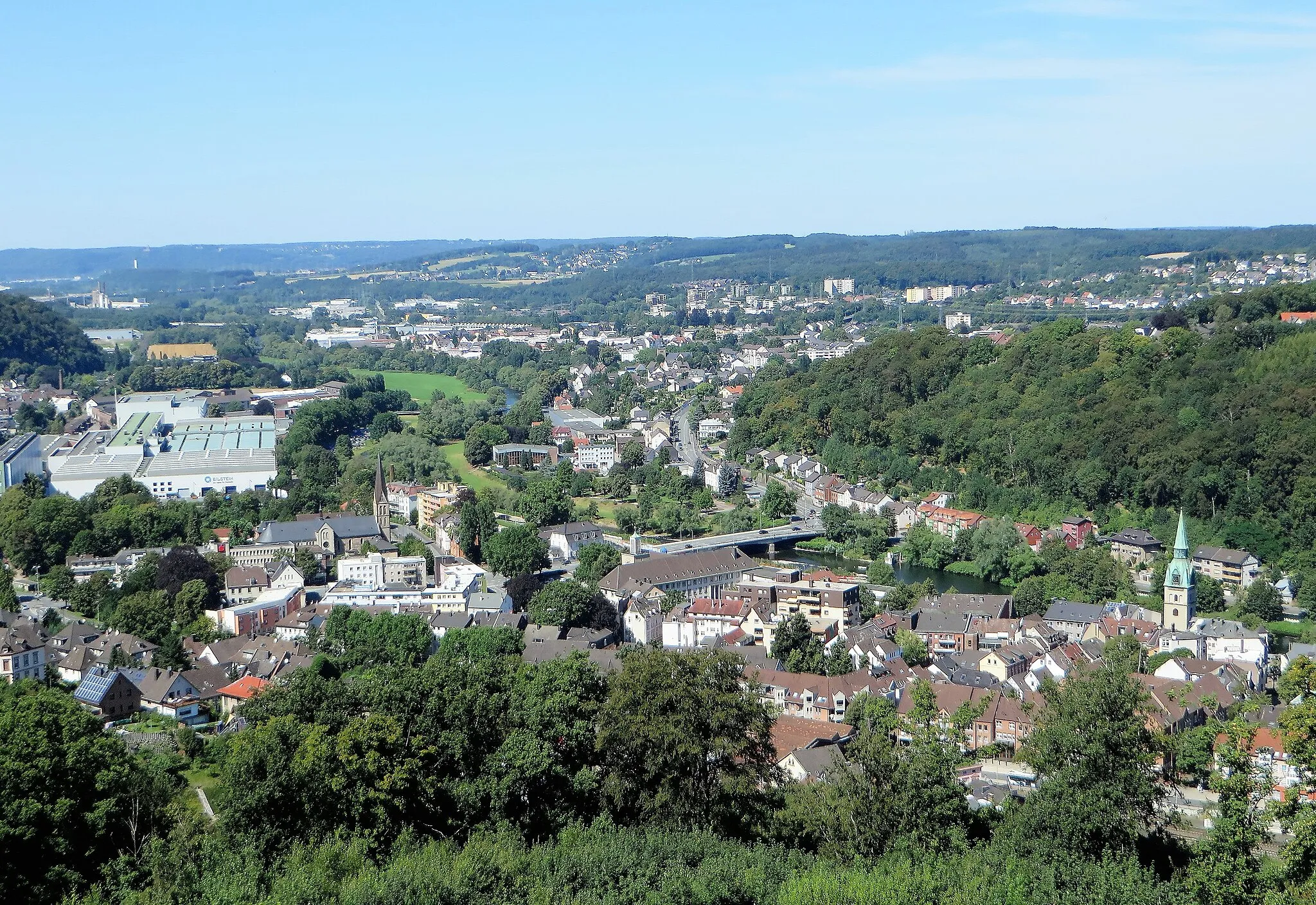 Photo showing: Blick von Schloss Hohenlimburg auf den Hagener Stadtteil Hohenlimburg. Auf dem Foto von rechts nach links, Reformierte Kirche, Lenne mit Stennertbrücke und Lennepark, Rathaus, katholische Bonifatius Kirche und Kaltwalzwerk Bilstein. Rechts der Waldhang mit dem Möllerdenkmal liegt im Naturschutzgebiet „Steltenberg“. Im Hintergrund sieht man den Ortsteil Elsey und am Horizont in der Mitte den Stadtteil Berchum.