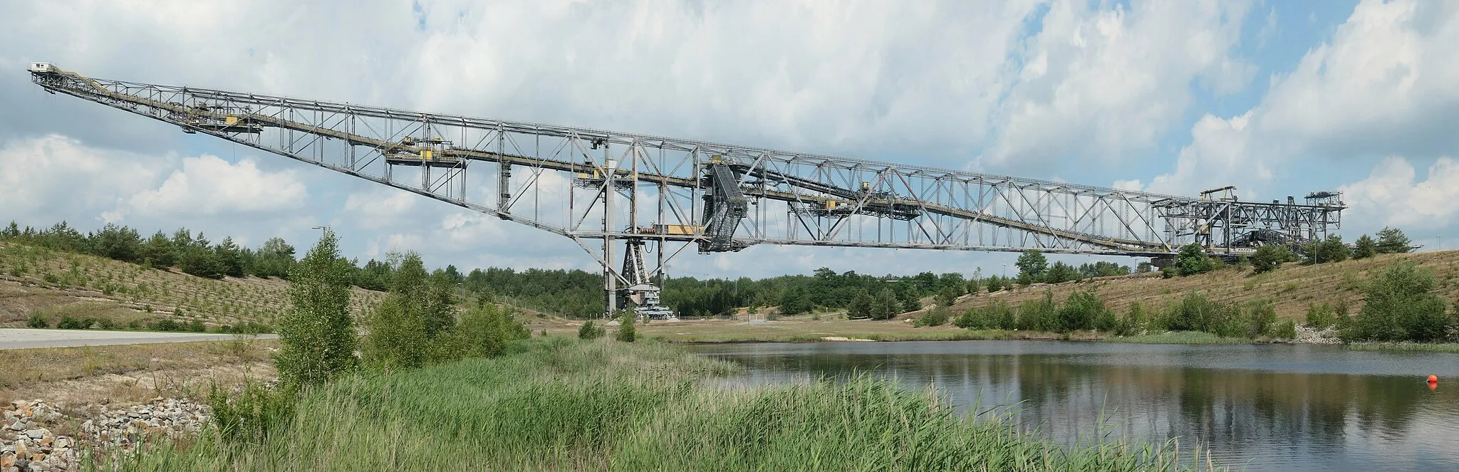 Photo showing: The overburden conveyor bridge transports and dumps the overburden supplied by two excavators to the opposite side of an open pit mine. The overburden capacity of the two excavators in the high and low cuts totals 60 metres, hence the name F60. The one pictured is the last of five F60 bridges built. With a length of 502 metres, a width of 204 metres, a height of 80 metres and a weight of 11,000 tonnes, they are the largest mobile technical working machines in the world.