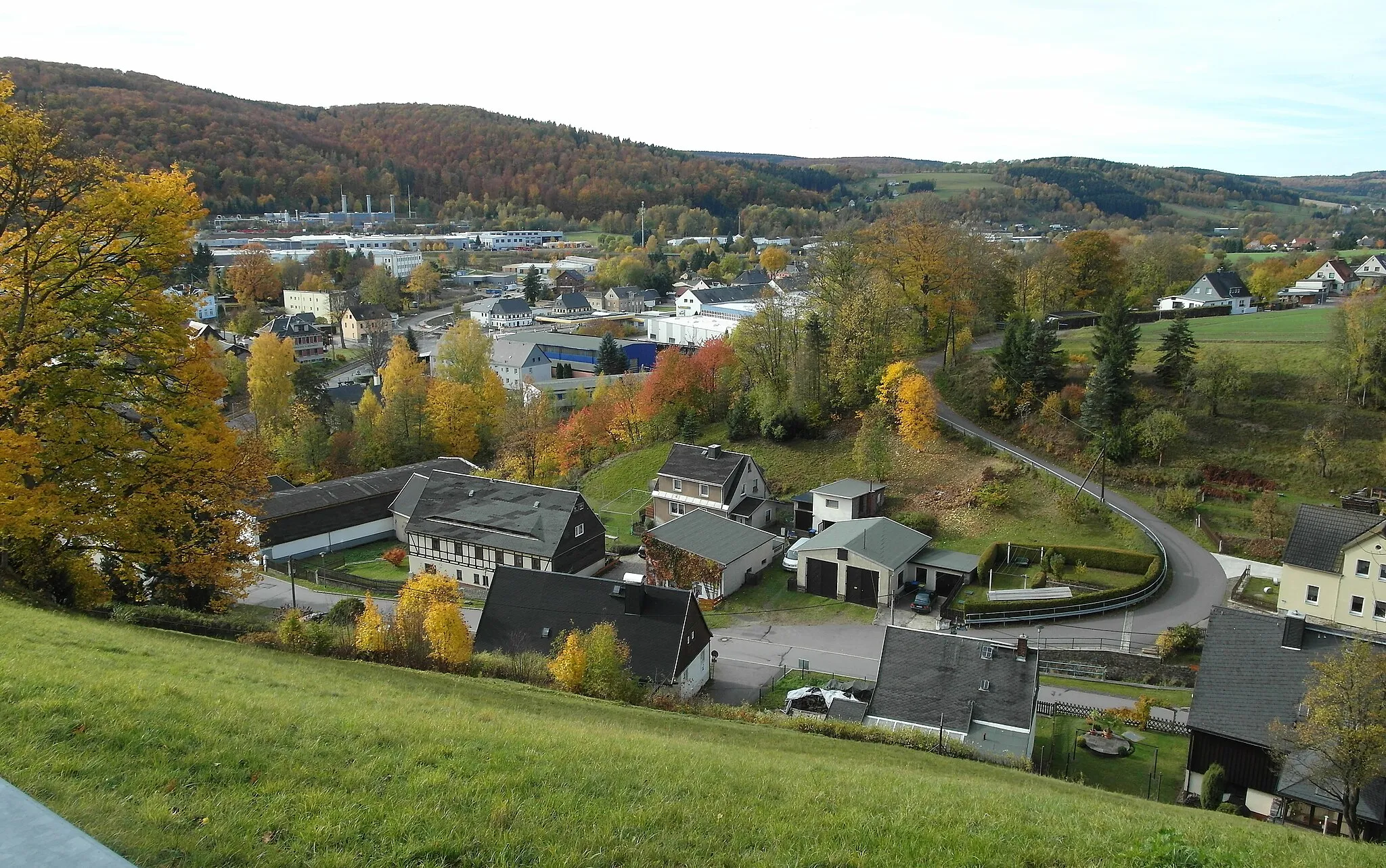 Photo showing: 30.10.2023  09526 Olbernhau-Oberneuschönberg / Grünthal: Blick von der Exulantenkirche (GMP: 50.652064,13.370481) nach Südwesten auf den Ortsteil Olbernhau-Grünthal. Im Vordergrund rechts windet sich die Straße Am Preißlerberg nach oben. In Bildmitte die einstigen Werkanlagen des VEB Blechwalzwerk Olbernhau (GMP: 50.651280,13.365830), heute von verschiedenen Firmen genutzt. Leicht links der Bildmitte steht in rostroten Klinkern erbaut das Bahnhofsgebäude Olbernhau-Grünthal, Grünthaler Straße 220 (GMP: 50.649830,13.364240). Es ist der heutige Endpunkt der Flöhatalbahn von Chemnitz - Flöha - Pockau - Olbernhau (Grünthal), einstmals bis Neuhausen führend. Links im Hintergrund, vor den Waldflächen am Bruchberg, sieht man am Heidenweg 34 die 3 Schornsteine der  
Gasverdichterstation Olbernhau (GMP: 50.645500,13.359500) und im Umfeld weitere Gewerbebauten. Am Horizont leicht rechts der Bildmitte zieht sich die Baumreihe vom Rübenauer Weg (GMP: 50.645230.13.340240) über die Hochfläche. Der Stadtkern von Olbernhau ist hier nicht mit im Bild.                                                                                 [SAM0343.JPG]20231030020DR.JPG(c)Blobelt