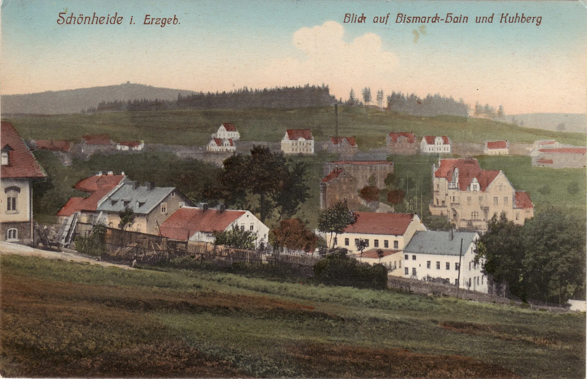 Photo showing: Fuchswinkel, ein Ortsteil von Schönheide im Erzgebirge: Seit um 1900 betrieb ein Bismarck-Komitee Schönheide die Errichtung eines Bismarck-Denkmals auch in Schönheide. 1910 kaufte die Gemeinde Schönheide die Waldfläche um den Fuchsstein (721, 9 Meter), gestaltet sie parkartig und nennt sie Bismarckhain. Zum 100. Geburtstag Bismarcks am 1. April 1915, mitten im Ersten Weltkrieg, wird ein Bismarck-Relief enthüllt. Unmittelbar nach dem Zweiten Weltkrieg entfernt die Gemeinde Schönheide das Relief und gibt dem Fuchsstein seinen Namen zurück. Blick vom Berg Knock (725,5 Meter) in Richtung Fuchsstein (721,9 Meter) und Kuhberg. Links am Rand das Anwesen des Bauern Rudert (Wohnhaus angeschnitten), unterhalb die Häuserzeile der Lindenstraße (beides Teile des Ortsteils Baumannsberg. In der Bildmitte mit Schornstein die nach 1990 abgerissene Textil-Druckfabrik Arlt (Hauptstraße, Ecke Schneeberger Straße), rechts davon die beiden Lenk-Villen (Hauptstraße 120 und 119 - vorn -), über dem Schornstein die Häuserzeile des Fuchswinkels mit den zum Bahnhof Schönheide Mitte führenden Gleisen der Schmalspurbahn Wilkau-Haßlau–Carlsfeld, darüber der Fuchsstein und links hinten der Kuhberg. Links am Rand angeschnitten das Haus des Bauern Rudert, rechts davon Häuser entlang der Lindenstraße,  alles schon zum Ortsteil Baumannsberg gehörend.