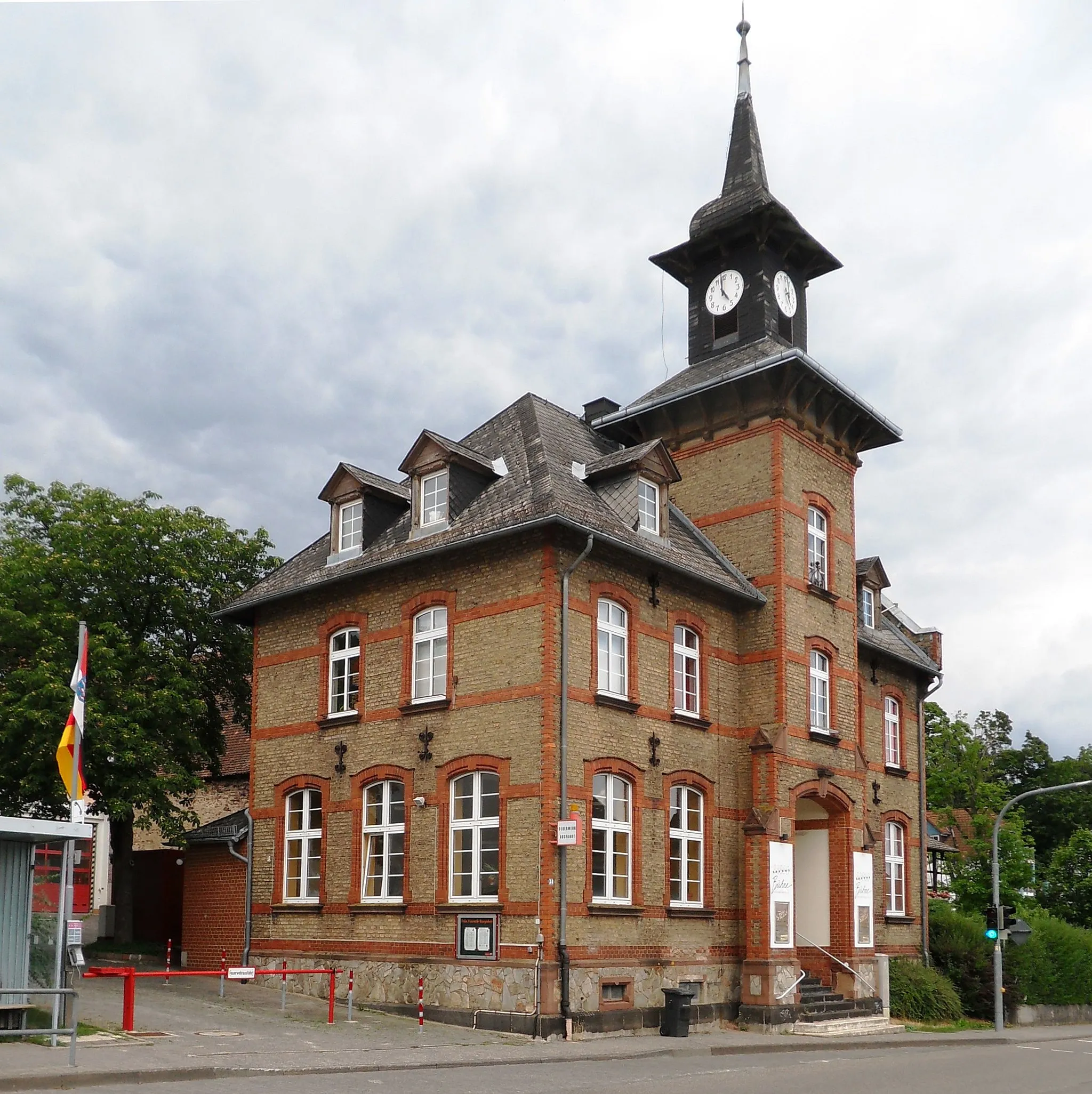 Photo showing: Blick von Westen auf die ehemalige Schule Mainstraße 34 in Georgenborn. Der denkmalgeschützte Backsteinbau mit einem kleinen Uhrturm in der Mittelachse wurde um 1900 in zentraler Lage an der Ortsdurchfahrt errichtet. Das für die kleine Gemeinde relativ aufwändige Schulhaus geht auf eine Stiftung des Barons Ferdinand von Krauskopf zurück