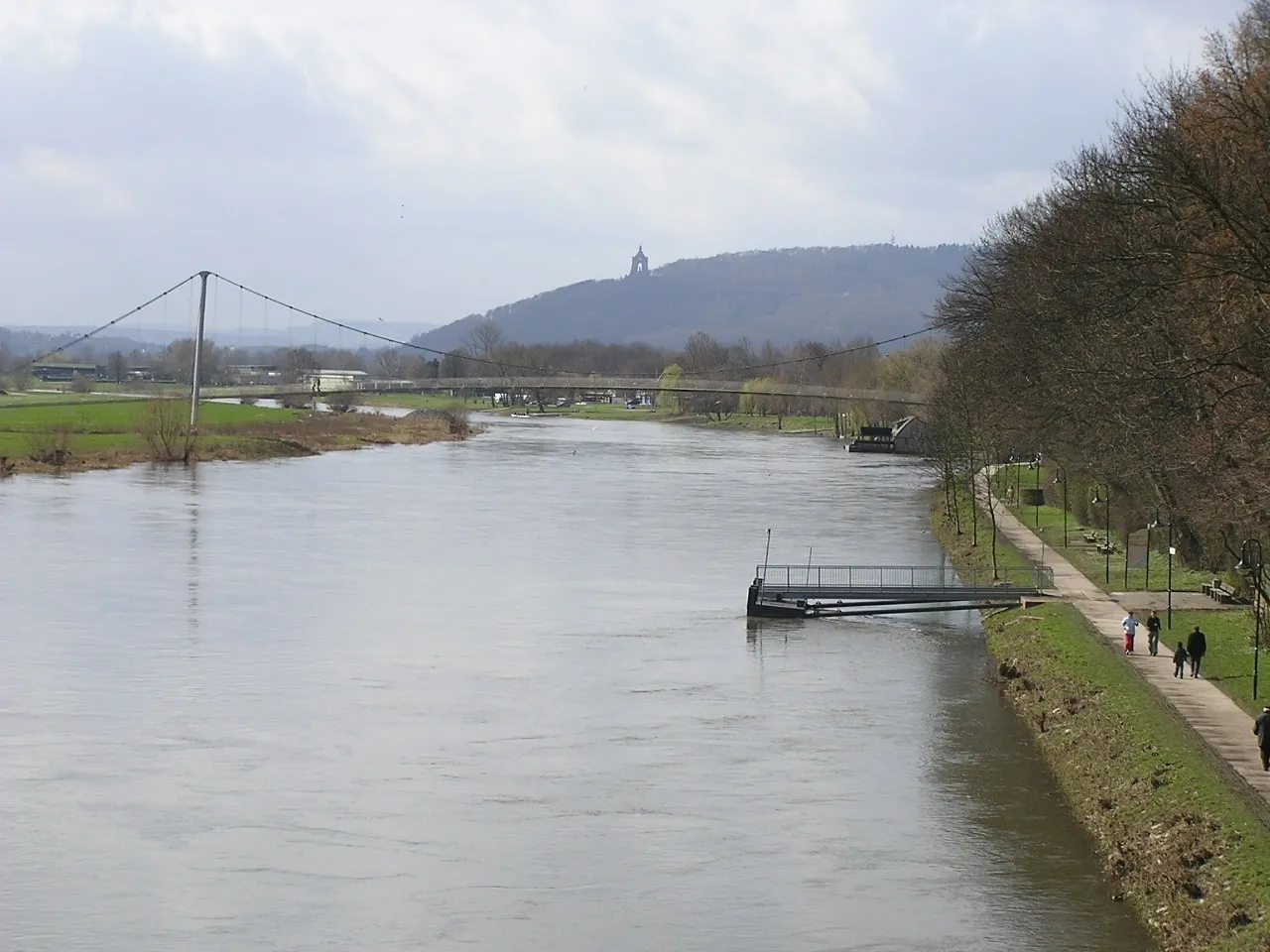 Photo showing: Blick vom westlichen Weserufer bei Minden (Westf.) in Nordrhein-Westfalen auf die Porta Westfalica im Süden. Links (außerhalb des Bildes) das Wesergebirge, rechts das Wiehengebirge. An dessen östlichem Abschluss das Kaiser-Wilhelm-Denkmal. Hinter der Hängebrücke am linken (vom Standpunkt rechten) Weserufer die Schiffmühle Minden an der Westfälischen Mühlenstraße und am Weserradweg.