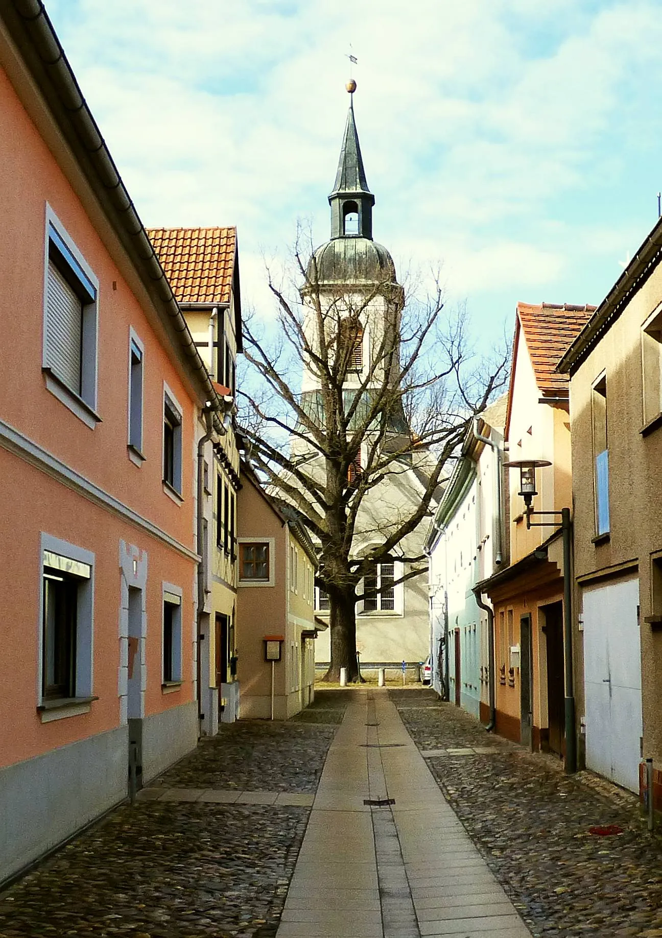 Photo showing: Die Kirchgasse in Ortrand. Im Hintergrund ist die Stadtkirche St. Barbara zusehen. Diese wurde im 16. Jahrhundert errichtet und nach einem Brand im Jahre 1707, der das Bauwerk schwer in Mitleidenschaft zog, unter dem Erbauer der Dresdner Frauenkirche George Bähr erneuert und mit dem heute zusehenden Turm versehen.