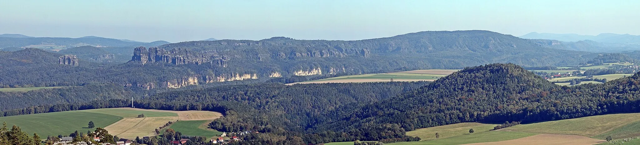 Photo showing: Saxon Switzerland: view from top of the Papststein (451 m) to the climbing peak Falkenstein (left, 381 m), the long group of rocks named Schrammsteine and the Großer Winterberg (right, 556 m).