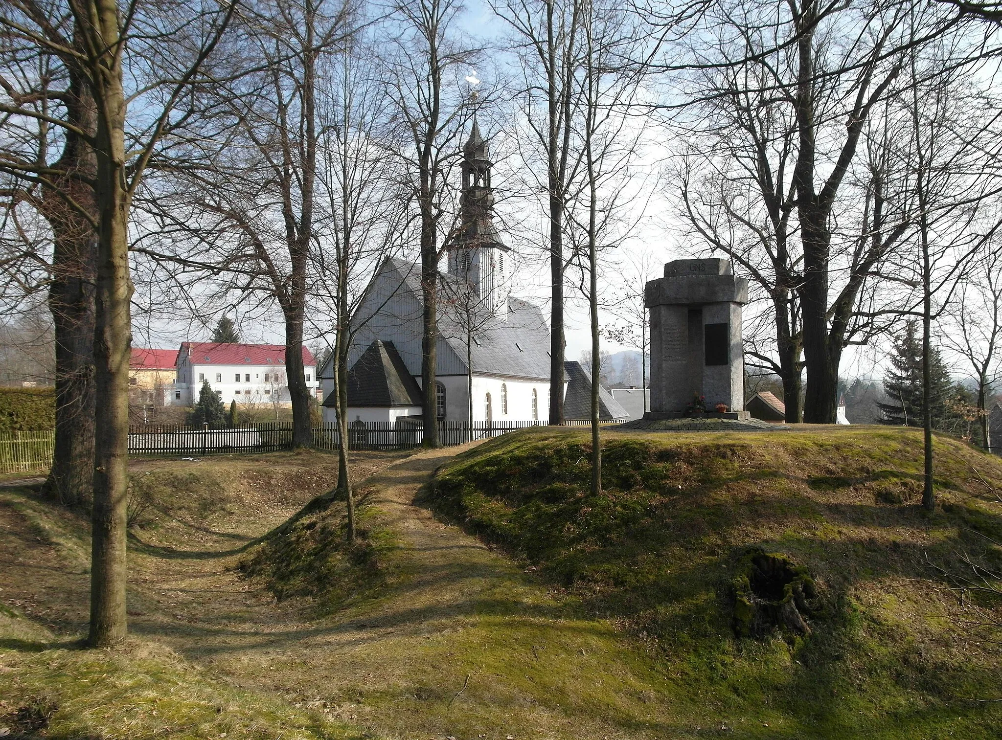 Photo showing: 09.03.2016     01848  Ulbersdorf (Oberulbersdorf, zu Hohnstein): Wasserburg am Schneckenberg (GMP: 50.952736,14.207554), dahinter die Kirche. Das 1432 als Olbersdorff erstmals urkundlich erwähnte Dorf, als Waldhufendorf in der zweiten Phase der deutschen Ostsiedlung angelegt. Ein Außenwall hinter der Kirche sowie ein Graben zeugen von einer mittelalterlichen, frühdeutschen Wasserburg am Schneckenberg mit Turmhügel aus der ersten Hälfte des 13. Jahrhunderts. Die Kirche entstand Ende des 17. Jahrhunderts als Saalkirche auf den Grundmauern des Vorgängerbaus. Sie erhielt 1699 einen hölzernen Dachreiter mit einer laternengekrönten, frühbarocken Haube. An der Südwand der Kirche sind sechs künstlerisch gestaltete Grabmäler der Ulbersdorfer Patronatsherrschaft aus den Jahren 1593 bis 1629 erhalten. Als ausführende 
Künstler sind unter anderem Michael Schwenke (1602) und dessen Sohn Hans (1611) belegt.                                                                                                     [SAM3511.JPG]20160309260DR.JPG(c)Blobelt