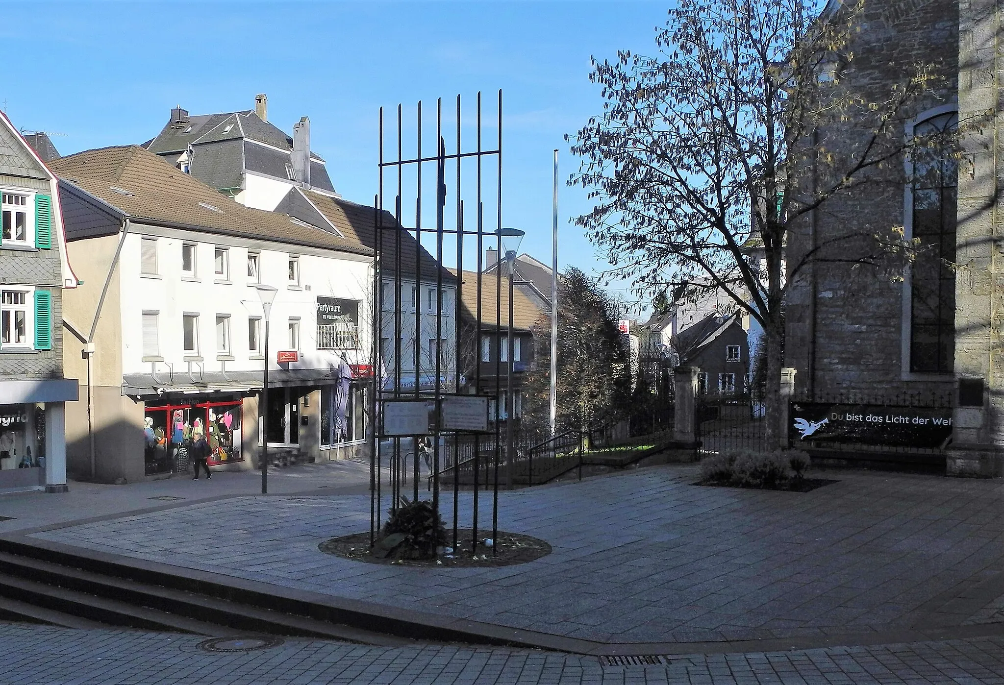 Photo showing: Mettmann, Lavalplatz, im Vordergrund die Gitterstäbe des Koburg-Mahnmals "Zum Gedenken an die Opfer der nationalsozialistischen Diktatur (1933-1945)", rechts im Schatten die evangelische Kirche, links von der Sonne bestrahlt das Wohn- und Geschäftshaus Freiheitstr. 20