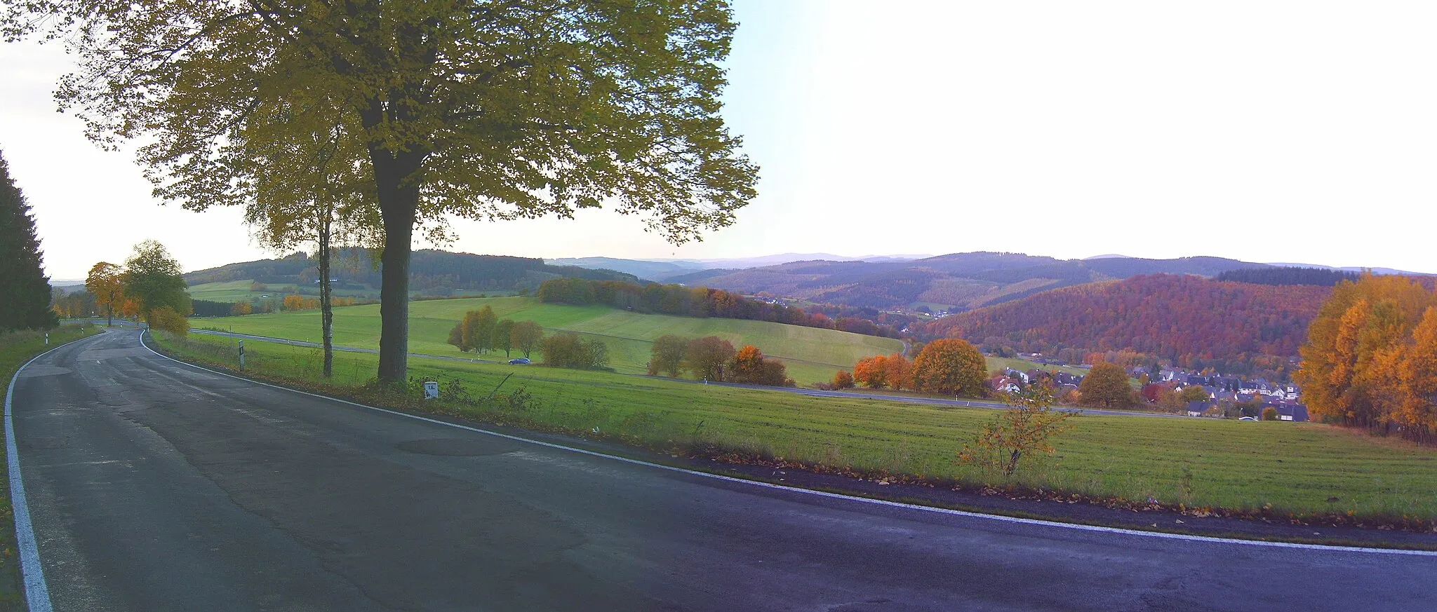 Photo showing: Siegerland-Landschaft von der Serpentine am Nordwestrand der Überquerung der Haincher Höhe im Südwesten des Rothaargebirges aus mit dem namensgebenden Ort Hainchen im Vordergrund. In zweiter Reihe die um eine Höhenstufe höheren Berge des westlichen Rothaargebirges: In der Mitte die Doppelkuppe aus Kindelsberg (618 m hoch, 21,6 km entfernt) und Martinshardt (616 m; 20,6 km), nach rechts übergehend in die Südwestkuppe (605 m; 24,0 km) und den eigentlichen Gipfel des Hohen Waldes (23,7 km; 655 m); halbrechts herauslugend die Alte Burg (633 m, Siegerländer Rothaar-Vorhöhen), rechts die Randhöhen des Massivs um die Oberste Henn (676 m). Vgl. auch Deutschle-Panoramen.